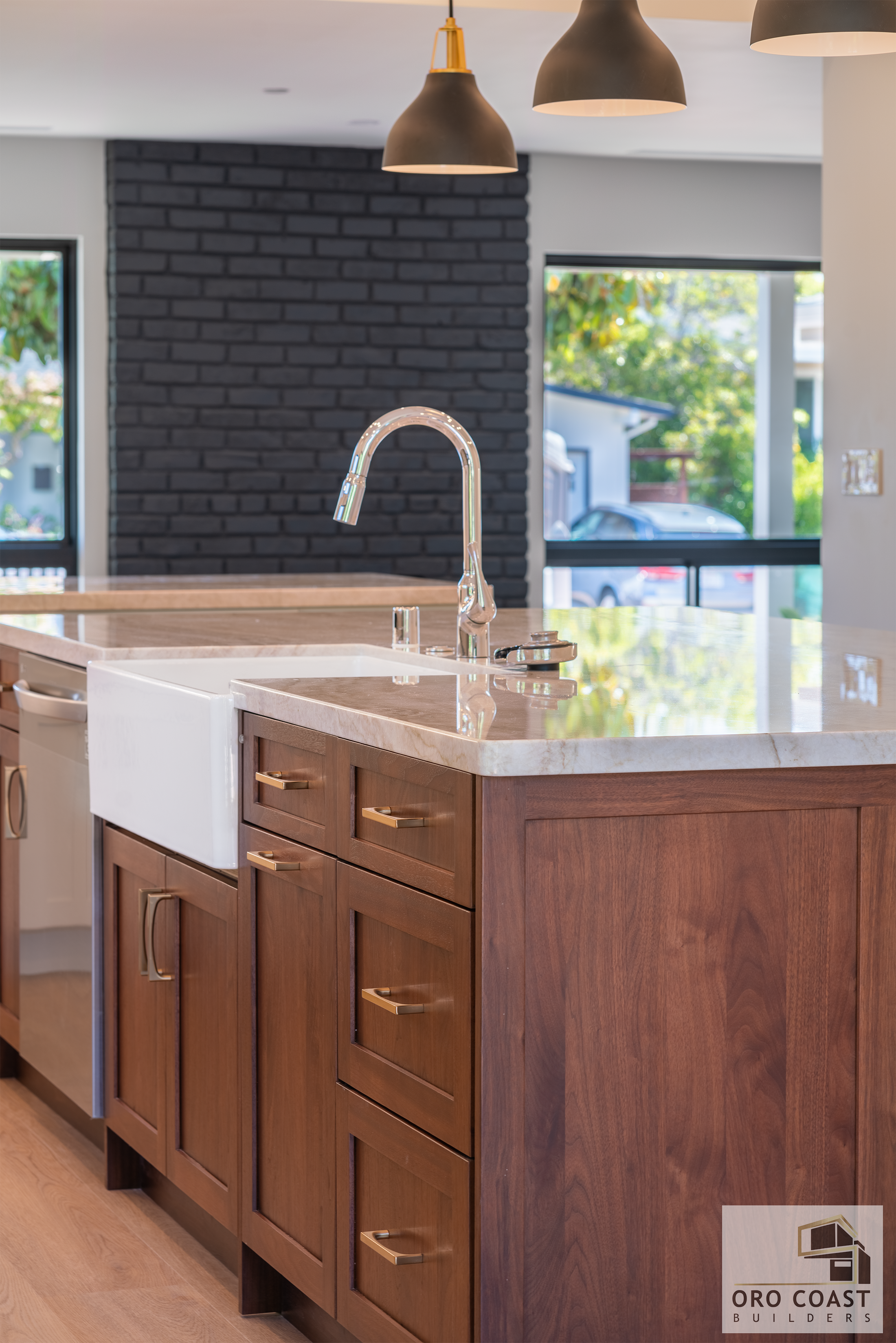 Modern kitchen with wooden cabinetry, marble countertops, a white farmhouse sink, a chrome faucet, and pendant lighting, with large windows showing a view of greenery outside.