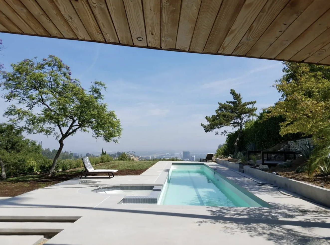 Modern backyard pool with lounge chairs, surrounded by trees and greenery, overlooking a cityscape under a blue sky.