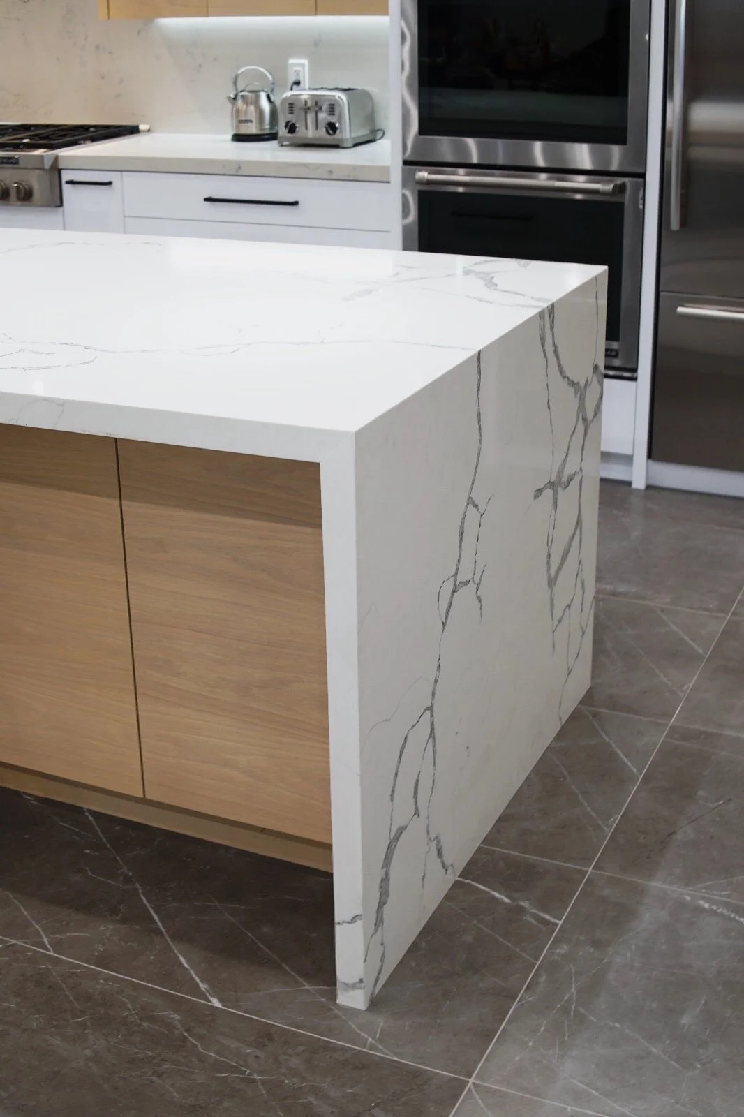 Close-up of a kitchen island with a white marble countertop with gray veining, and wooden cabinets below. The background shows a white counter with kitchen appliances, and to the right there's a stainless steel oven.