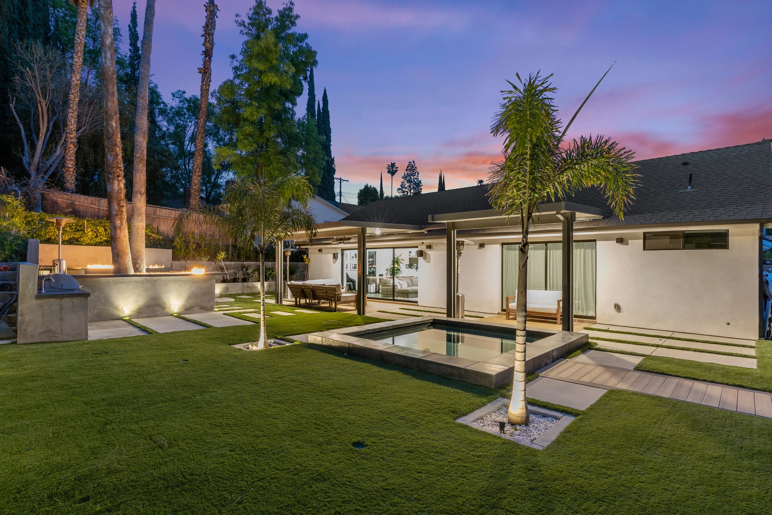 A modern backyard during sunset with a small pool, palm trees, outdoor seating, a fire feature, and a paved walkway leading to a house with large glass doors and windows.