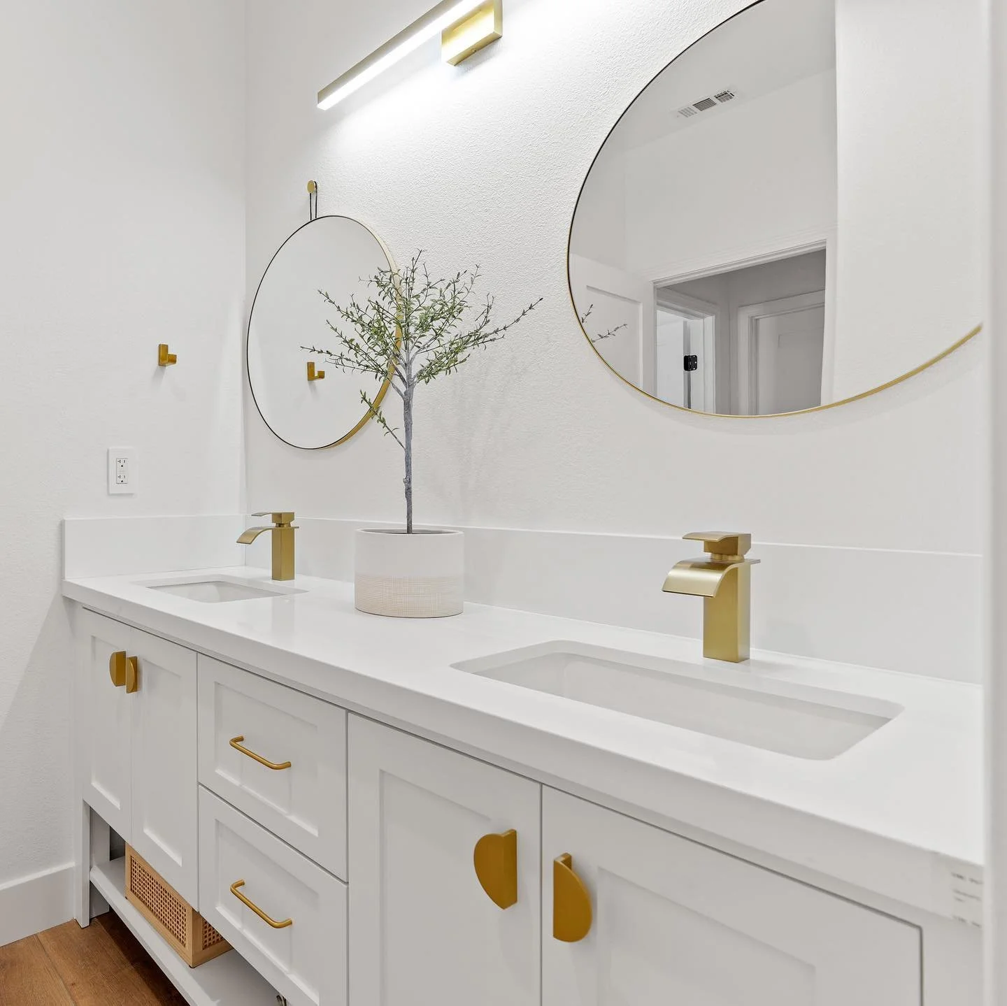 Bathroom vanity with double white sinks, gold faucets, gold cabinet knobs, a potted plant, and two round mirrors on a white wall.