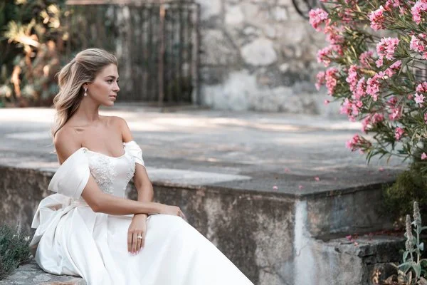 A woman in a white off-shoulder wedding dress sitting on a stone step next to a pink blooming bush.