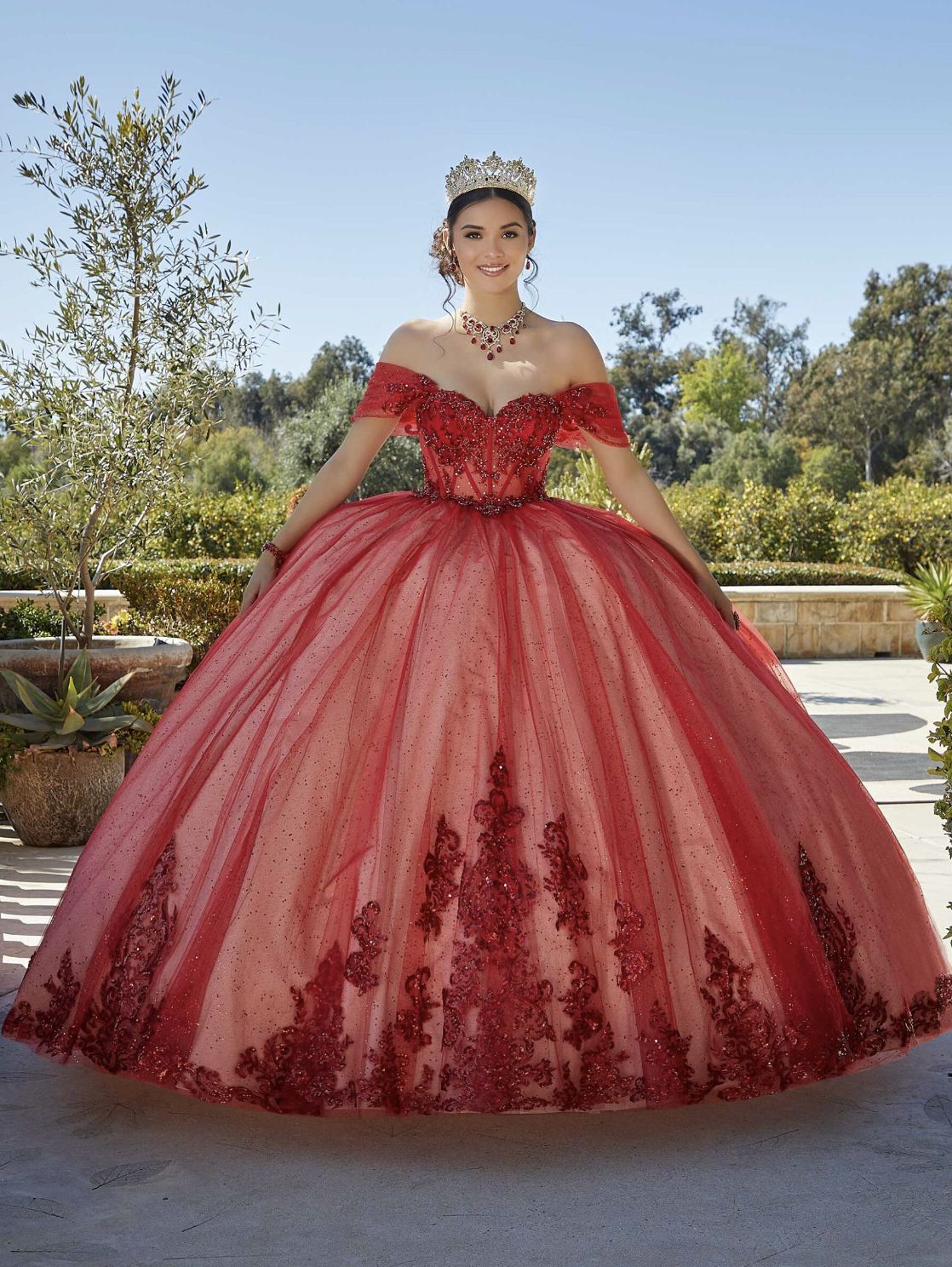 Young woman in an elaborate red ball gown with off-the-shoulder sleeves, lace and embroidery details, wearing a crystal crown and jewelry, standing outdoors in a garden with trees and plants.