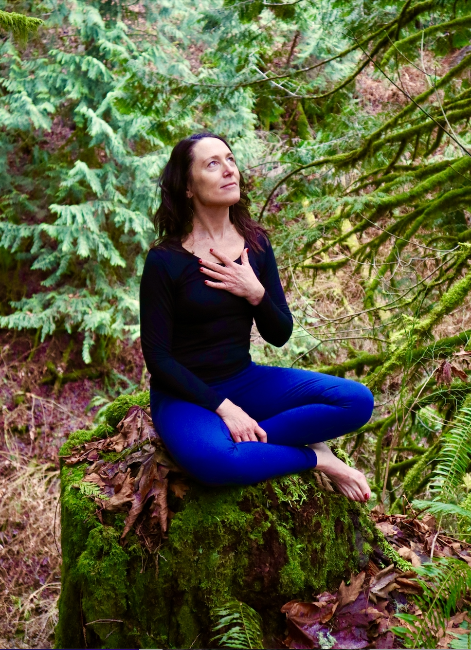 Cat Enright in woods sitting cross legged with hand over heart