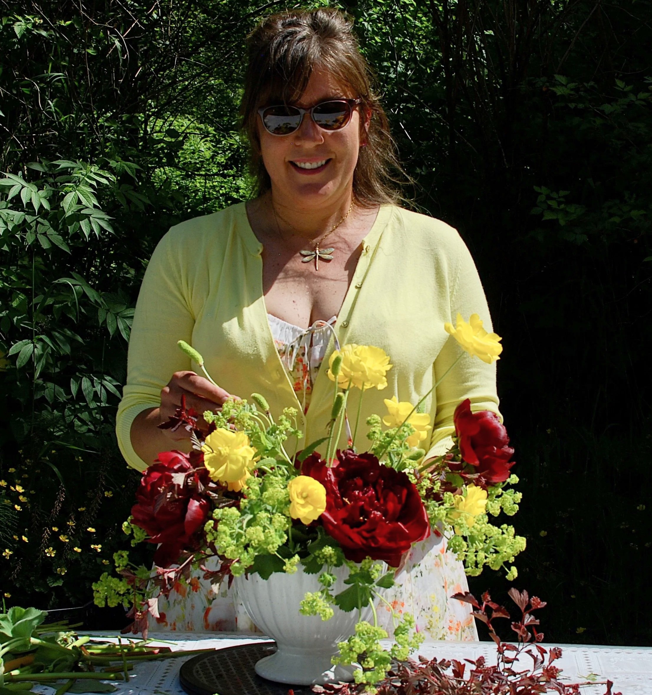 Young woman with dark hair tied in a bun, wearing glasses, hoop earrings, a blue sleeveless top, and a necklace, placing a bouquet of pink and white flowers on a table outdoors with green trees and a blue sky in the background.