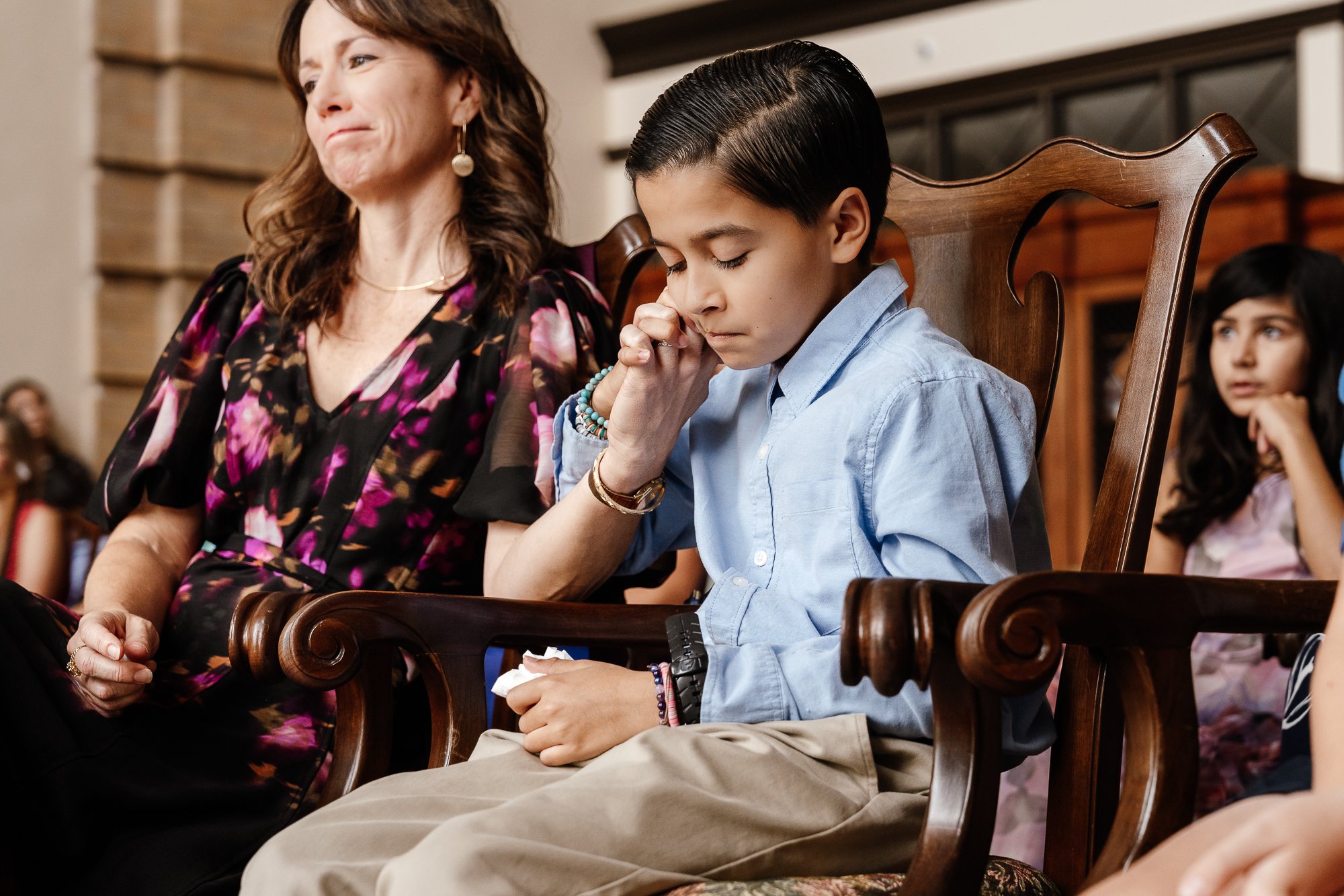 A young boy in a blue shirt sits on a wooden chair, holding a woman's hand, looking thoughtful. The woman, wearing a floral dress, is seated next to him, showing a slight smile. A girl is blurred in the background.