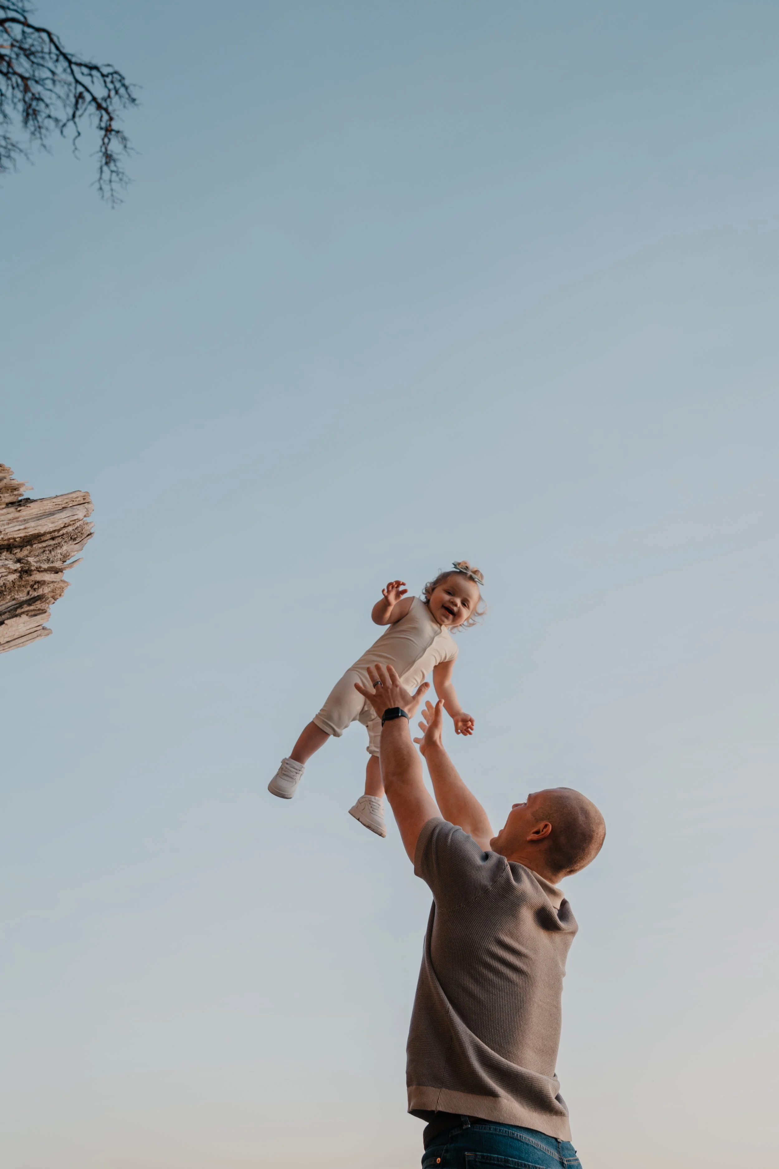 A man lifting a young girl into the air outdoors against a sky, with a tree on the left side of the image.