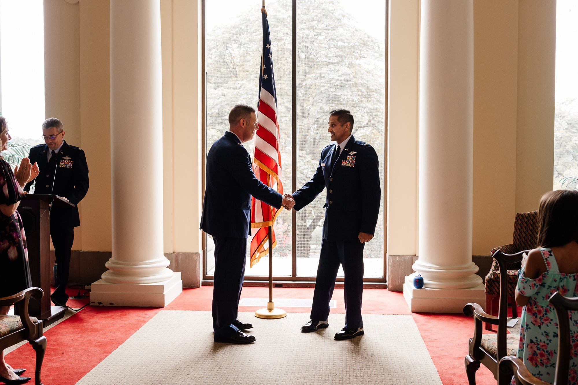 Military ceremony with two men shaking hands in front of an American flag, one in uniform. A small audience watches, with applause from a woman in the foreground. Large windows and columns suggest a formal indoor setting.
