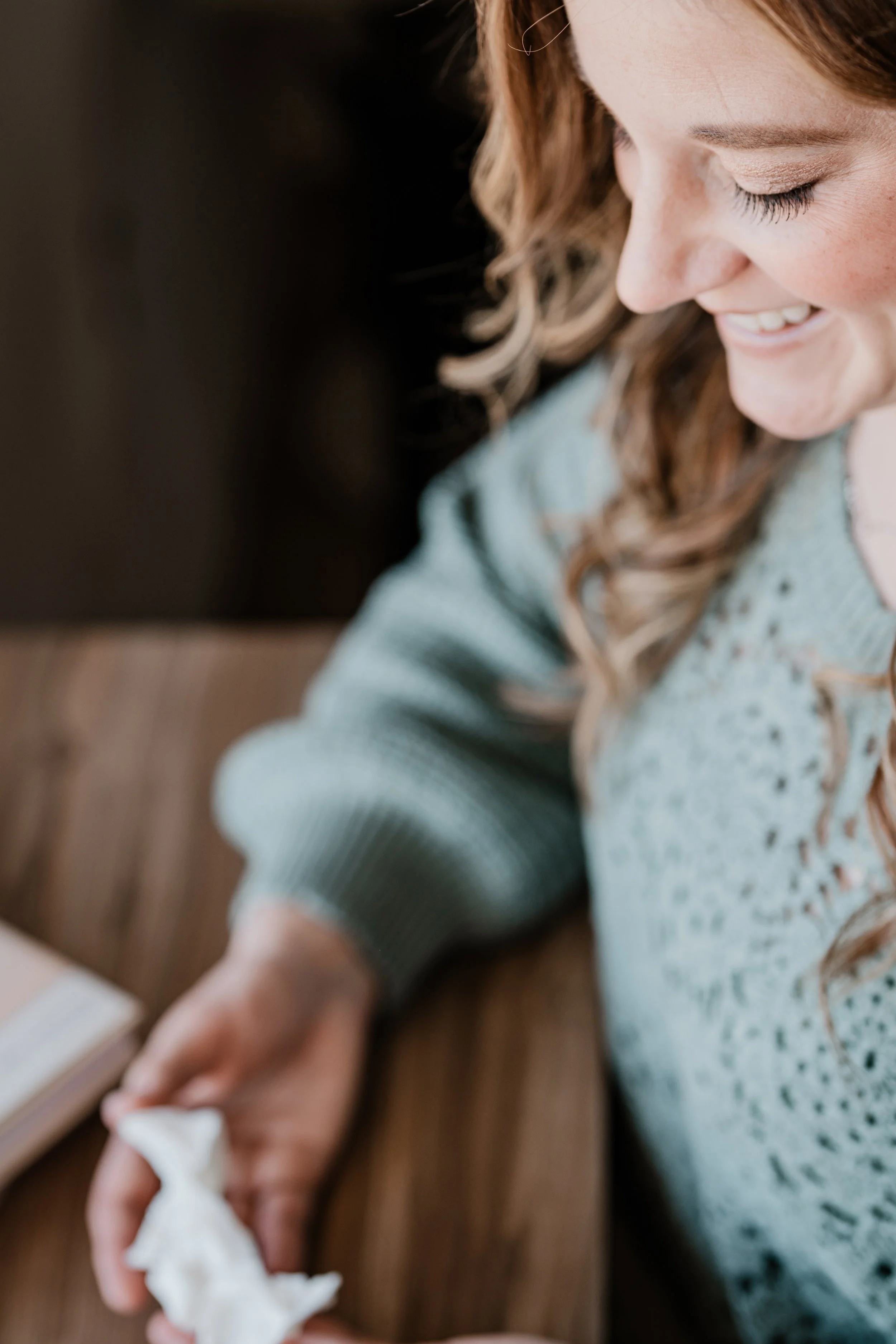 Close-up of a woman with long, curly brown hair smiling while holding a tissue in her hand, wearing a light blue sweater, sitting at a wooden table.