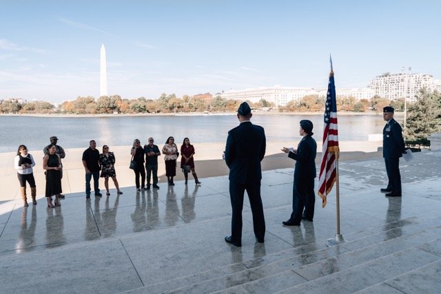 Military ceremony with individuals in uniform and civilians gathered near a river, American flag present, Washington Monument visible in the background.