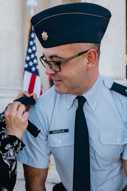 A uniformed individual in military attire, with glasses, identified by a name tag labeled "Castillo." The person is being assisted with their shoulder insignia as an American flag is visible in the background.