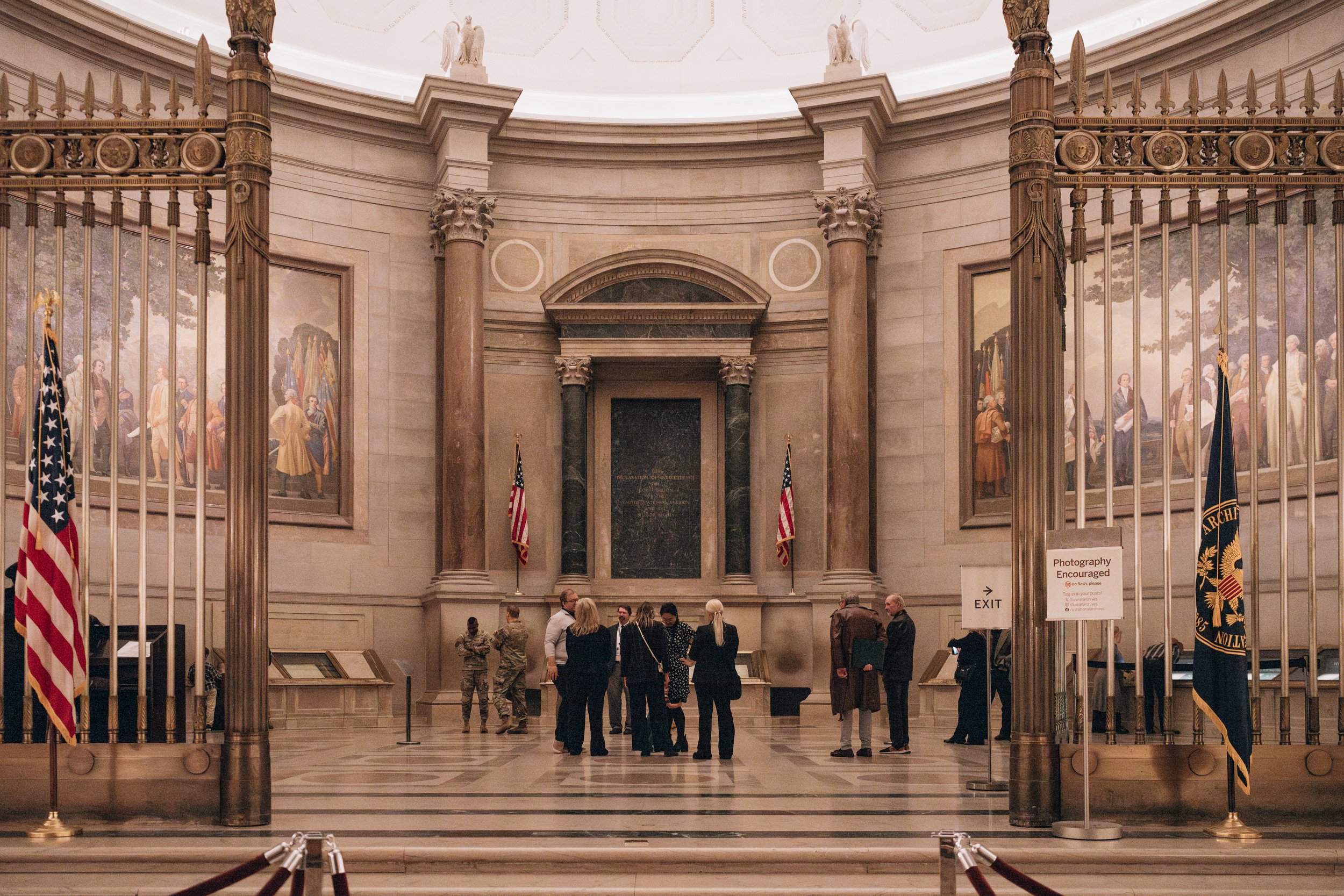 Interior of the National Archives Rotunda with people viewing historical documents, flanked by large American flags, with classical architecture and paintings on the walls.