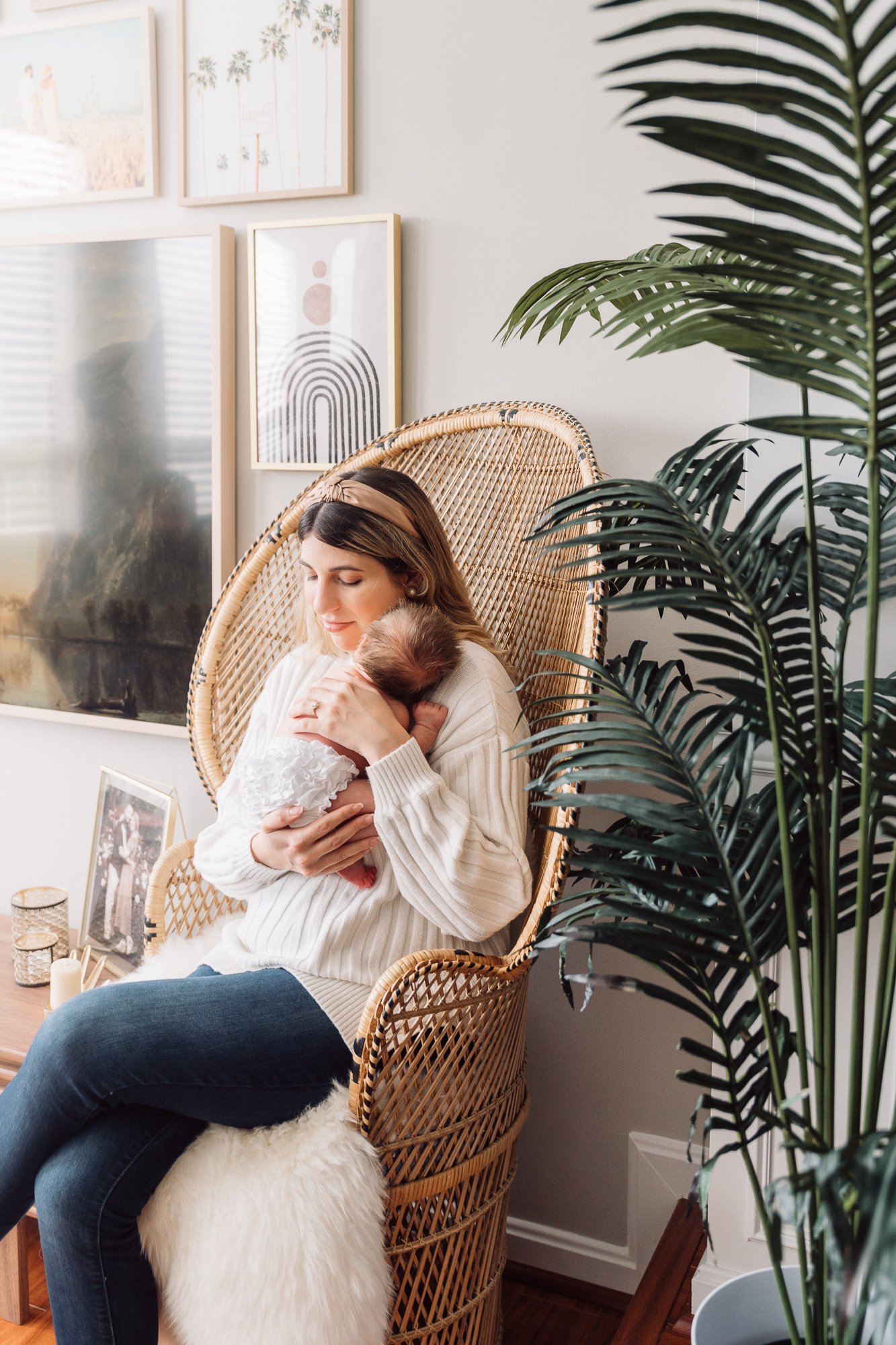 Woman sitting in a wicker chair holding a baby, surrounded by framed artwork and a large potted plant.