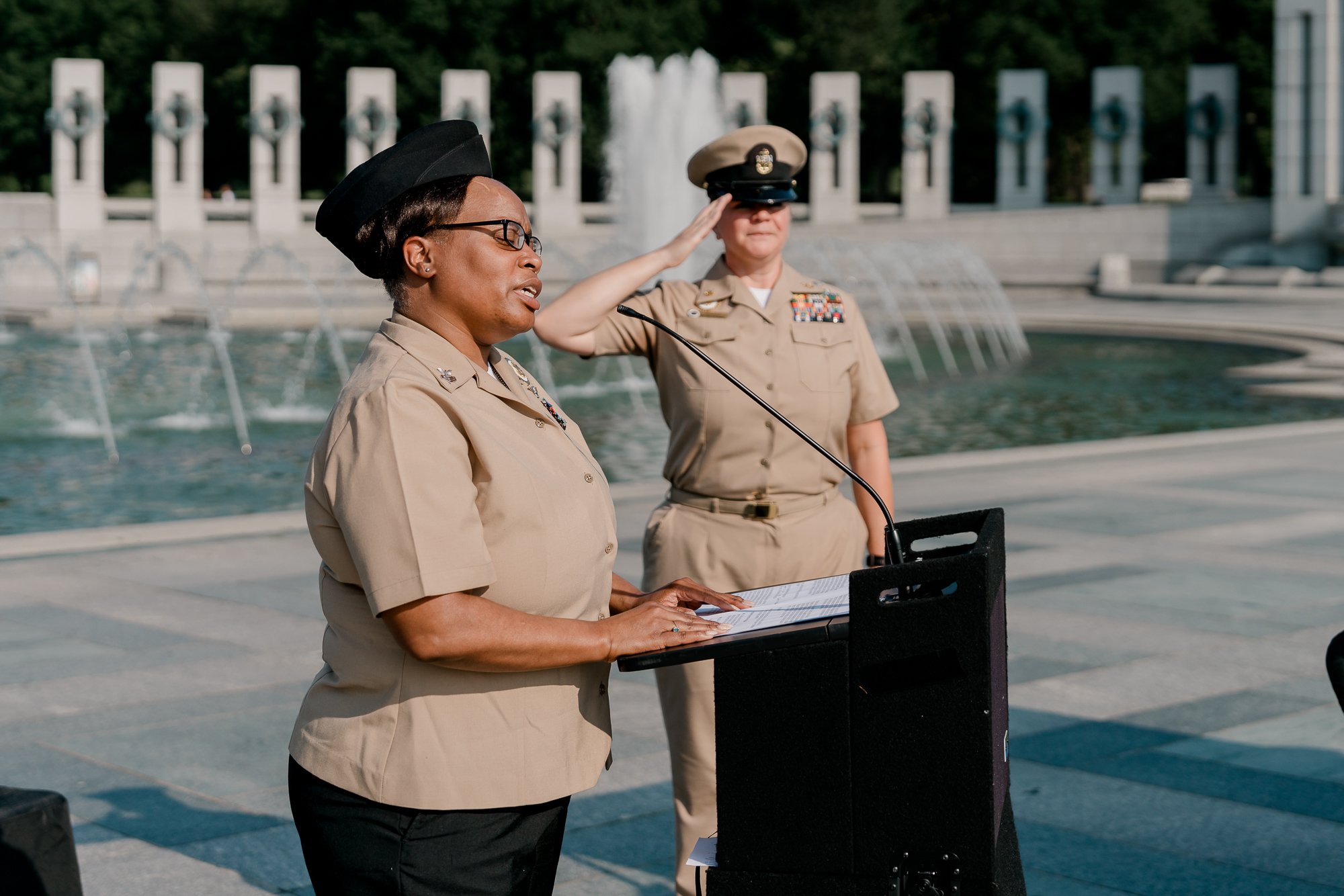Two military personnel in uniform, one speaking at a podium and the other saluting, in front of a fountain at a memorial site.