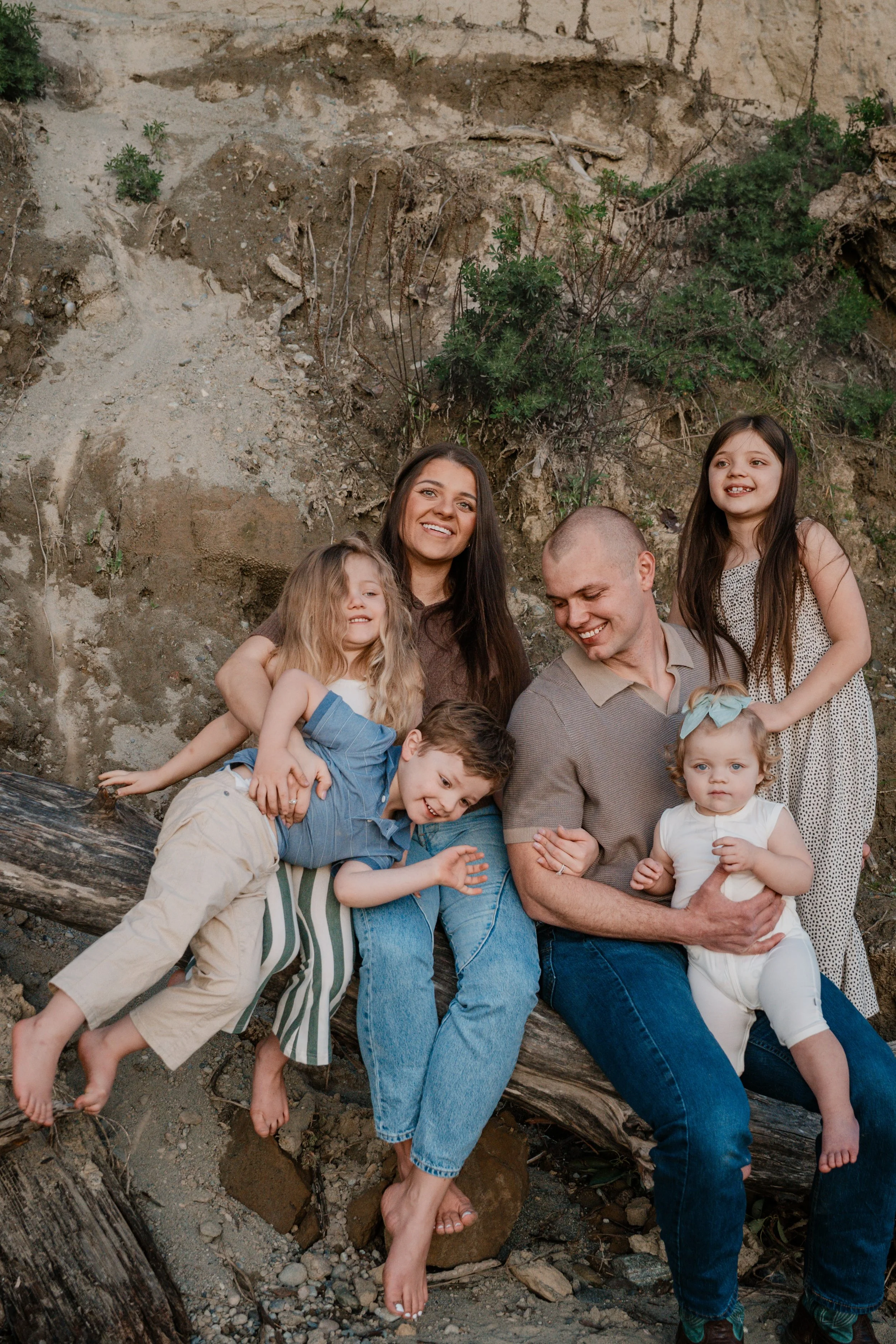 Family of seven sitting and lying on a fallen log against a rocky hillside with sparse vegetation.