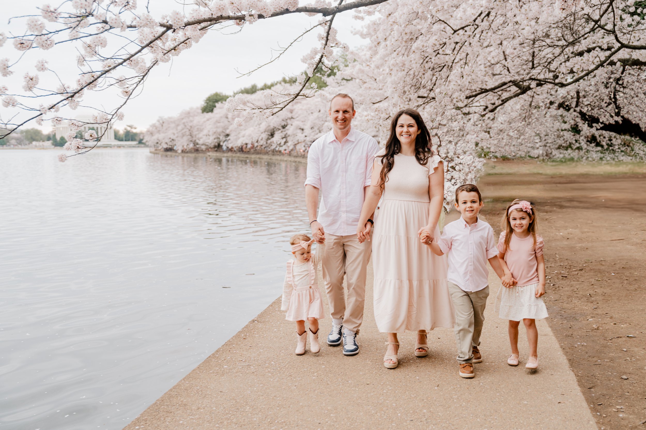 Family of five holding hands and walking along a lakeside trail with blooming cherry blossom trees in spring.