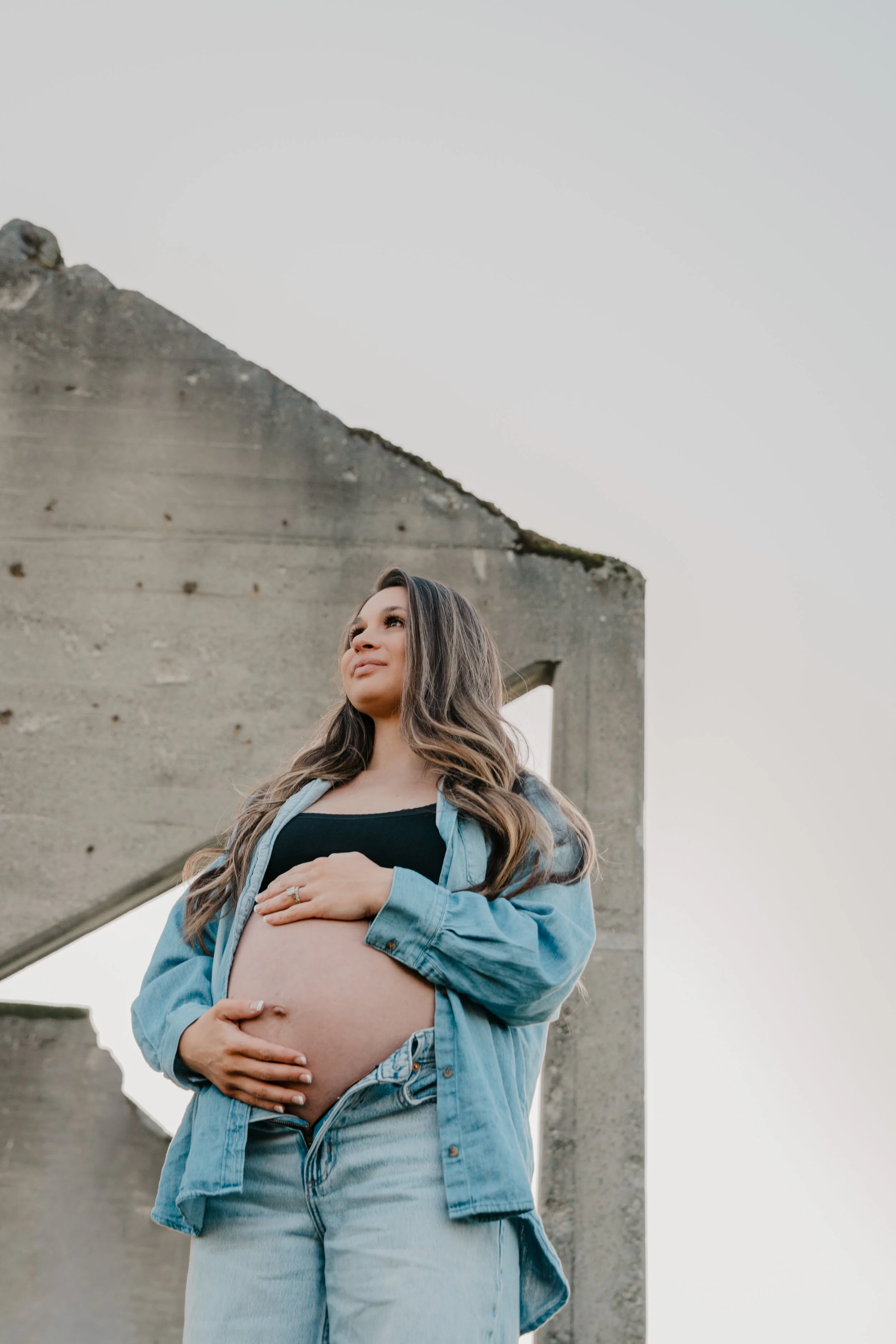 A pregnant woman with a bare belly, holding a bouquet of eucalyptus and white flowers, wearing a watch and denim jeans, standing next to a white curtain.