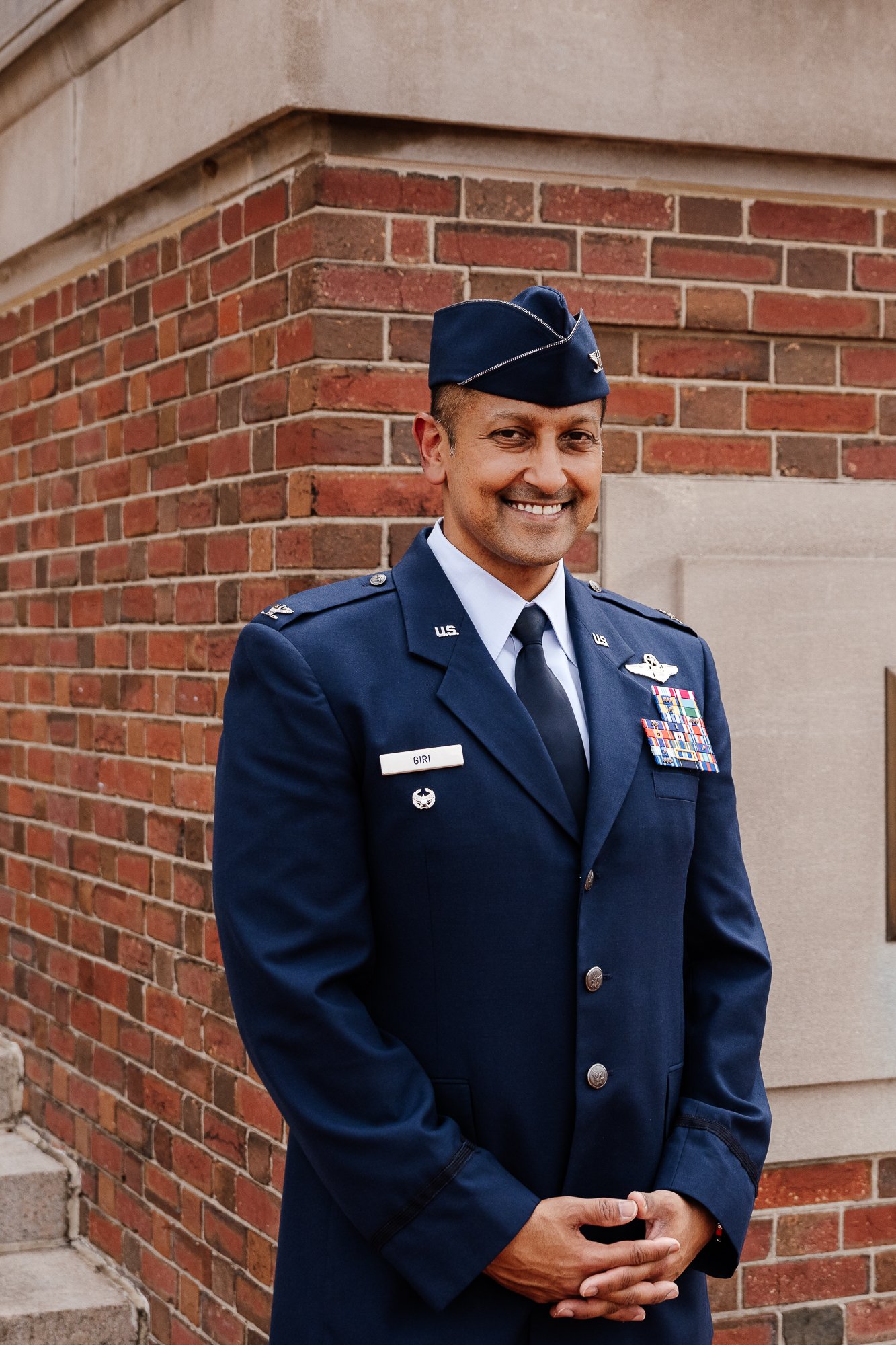 Air Force officer in blue uniform with ribbons, standing in front of a brick wall.