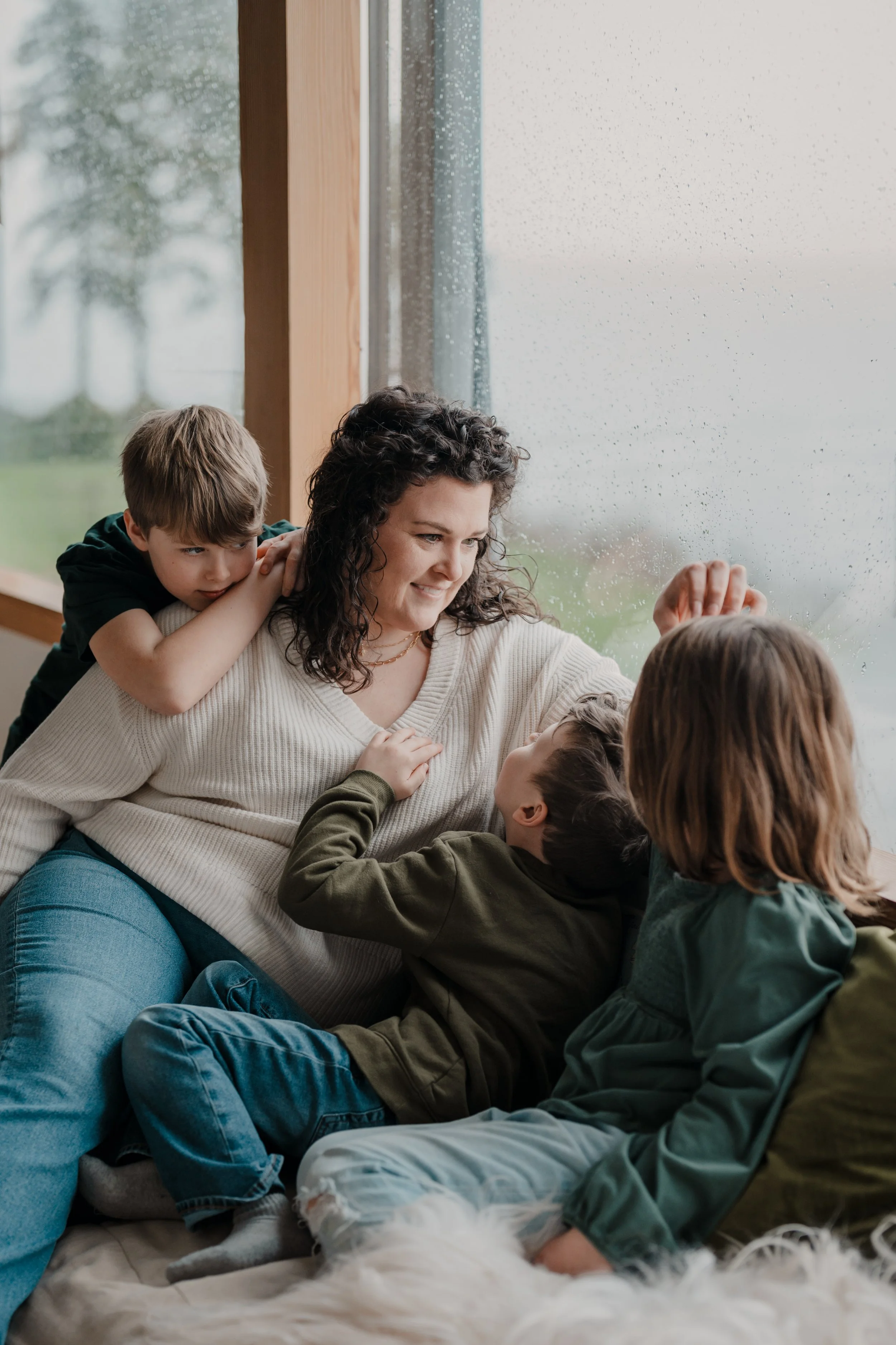 A woman with three children sitting by a rainy window, playing and cuddling with each other.