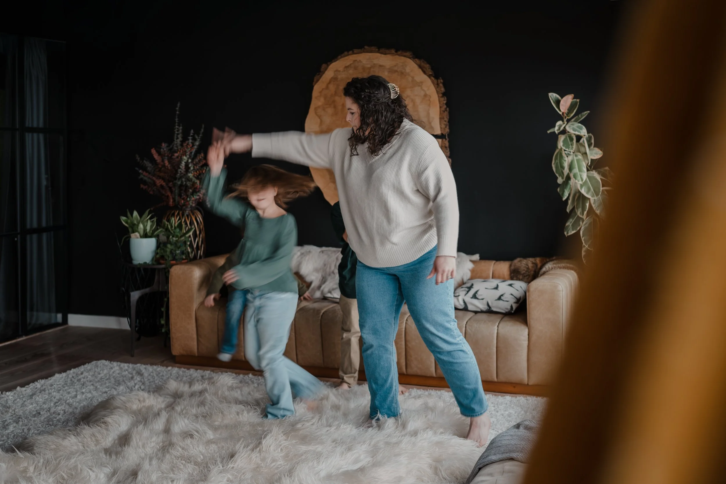 A woman and two children dancing in a living room with a fur rug, a brown sofa, and potted plants.