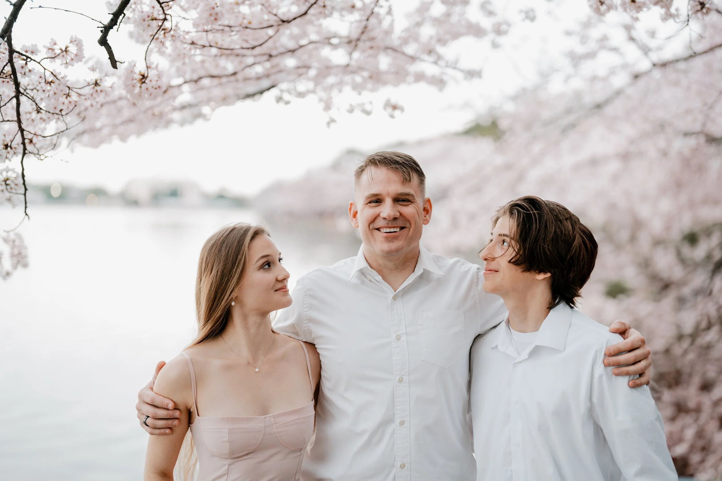 Three people standing outdoors by a lake with cherry blossom trees in bloom. The man in the middle is smiling, with his arms around a woman on each side. The woman on the left has long light brown hair and a necklace, while the person on the right ha