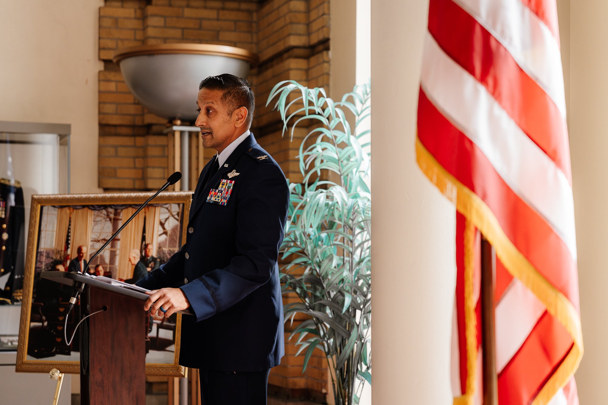 A military officer in dress uniform speaking at a podium, with an American flag and a framed photo in the background.
