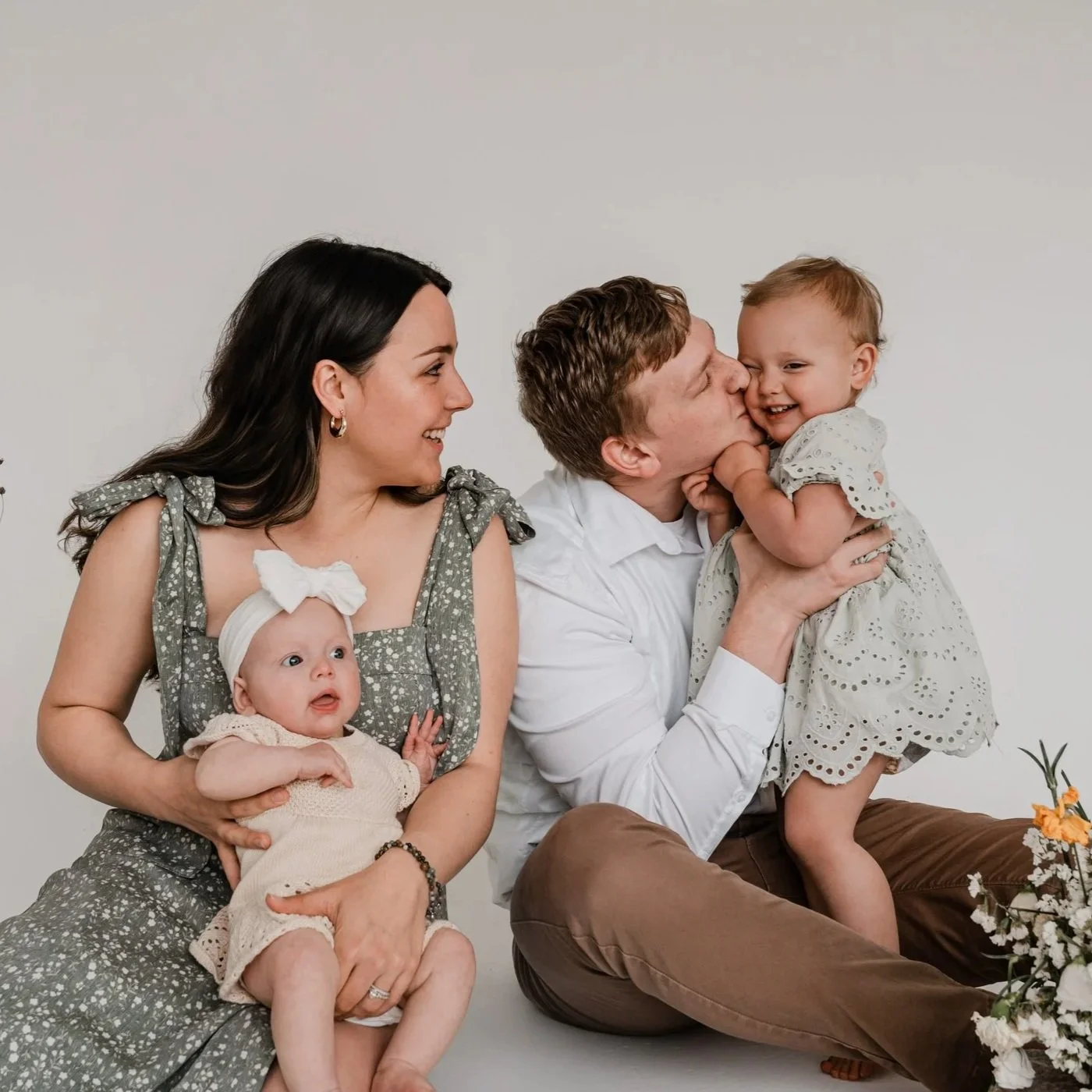 Family of four with two young children, one father kissing a young girl, mother holding a baby, in a studio with plain background.