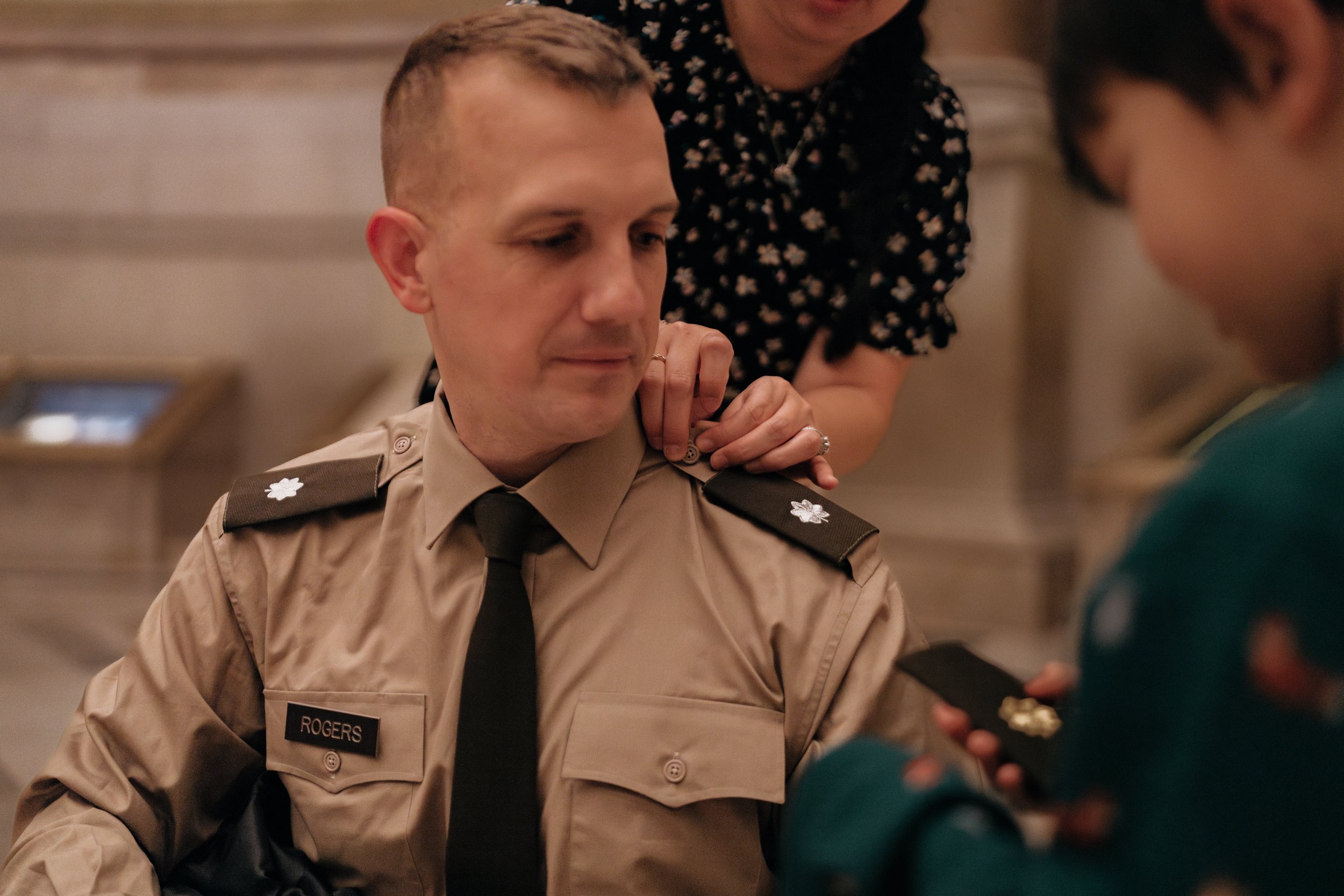 Person in a military uniform with name tag "Rogers," being pinned with an insignia on the shoulder by another individual. A third person is visible holding a patch.