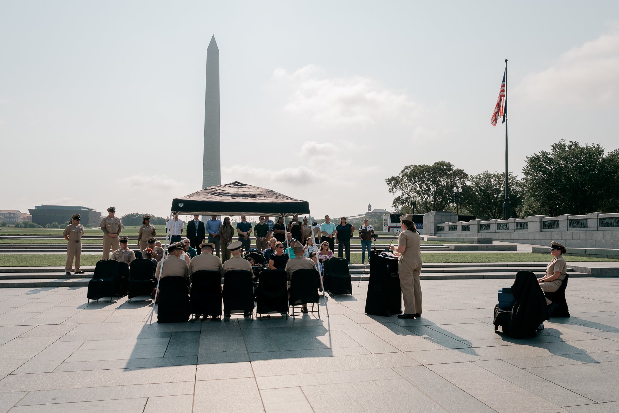 A group of people in uniform and civilians gathered for a ceremony at the National World War II Memorial with the Washington Monument and American flag in the background.