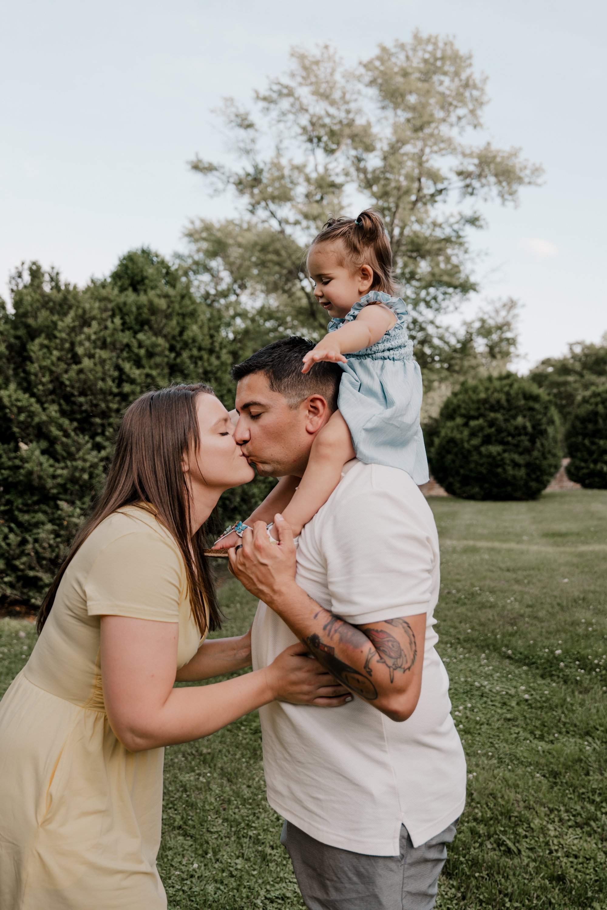 A family of three sharing a kiss outdoors, with a woman, man, and a young girl on a grassy area with trees in the background.