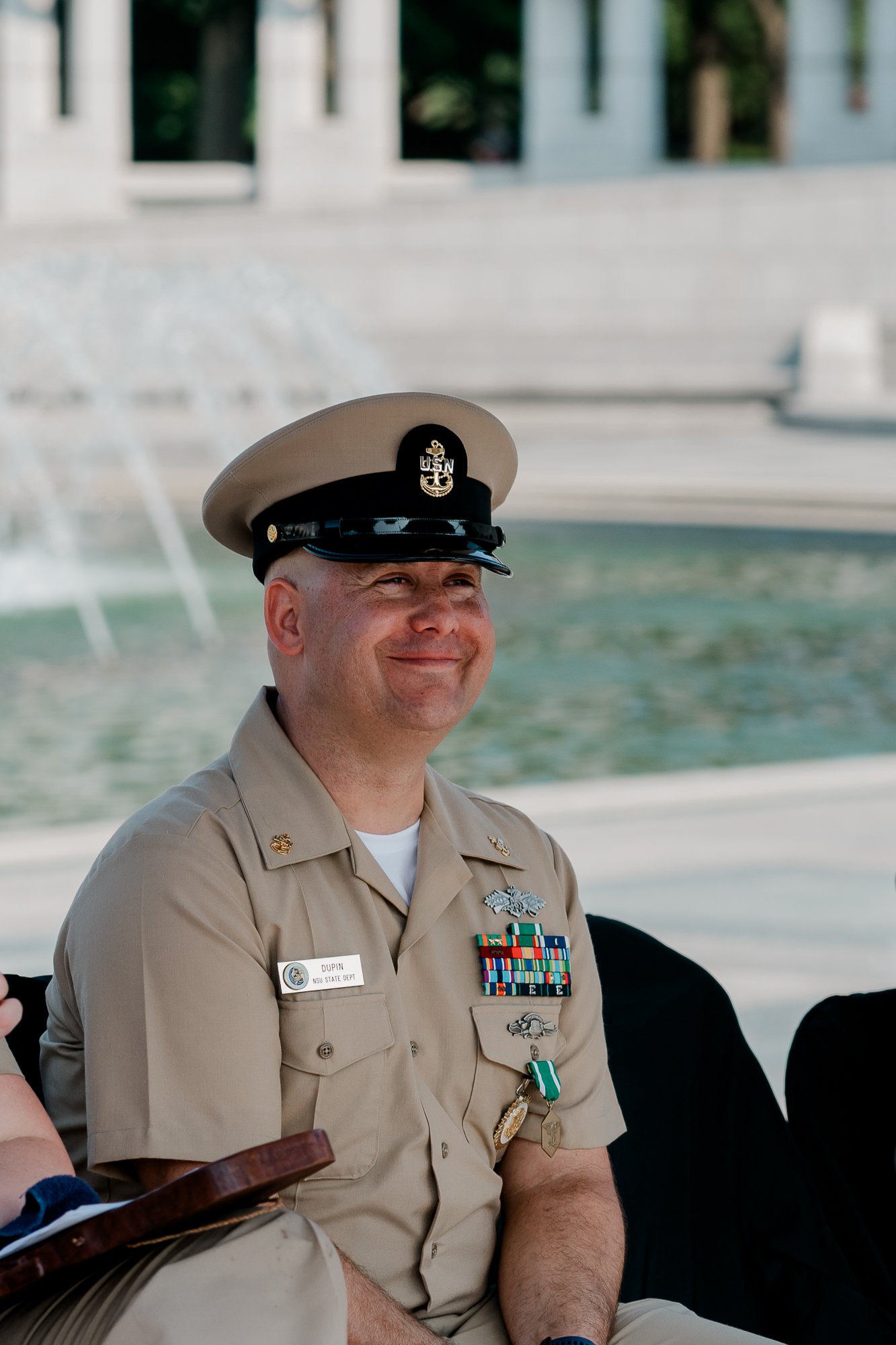 A person in military uniform with ribbons and medals, seated outdoors near a fountain.