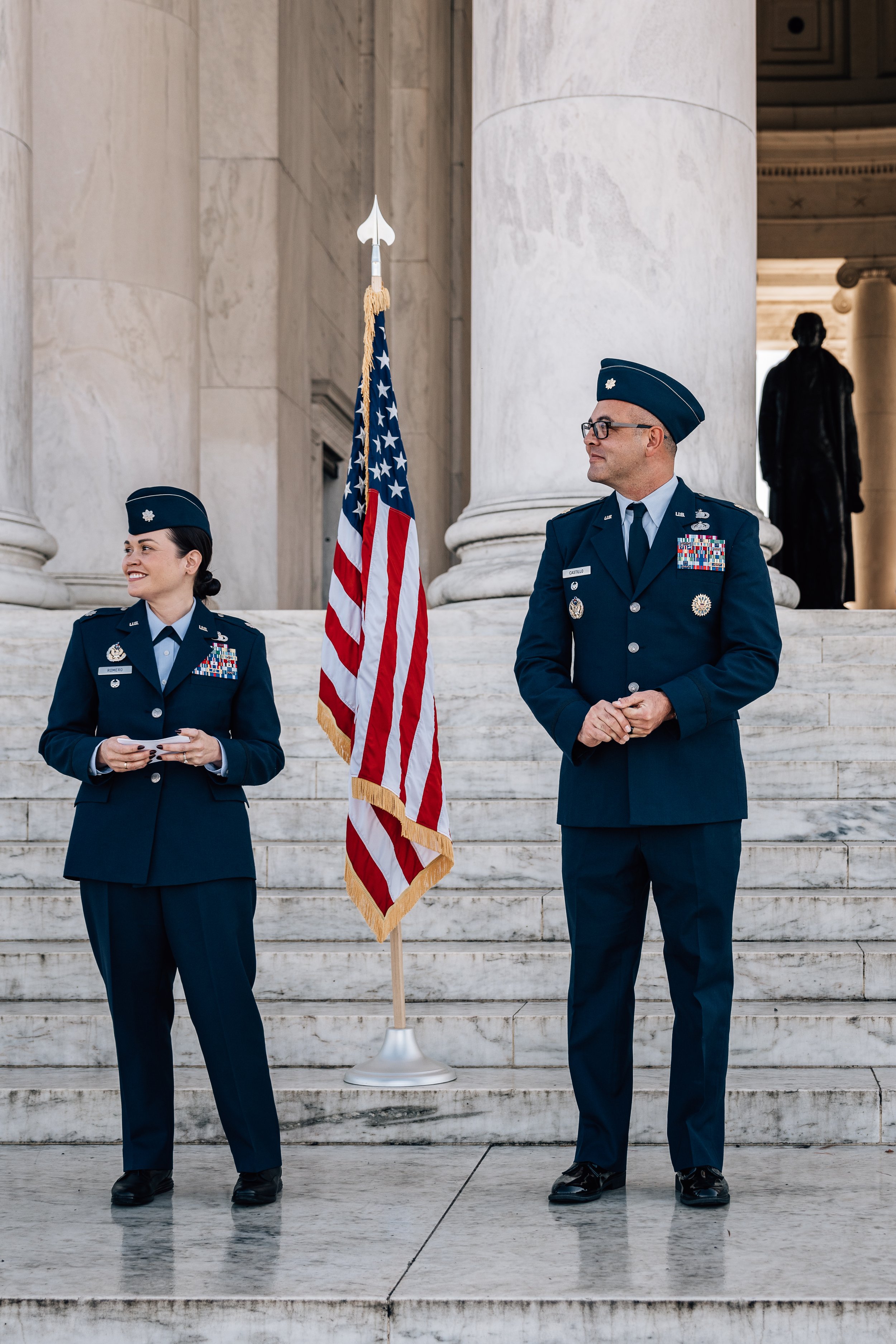 Two military personnel in dress uniforms stand next to an American flag on marble steps, in front of a large columned building.