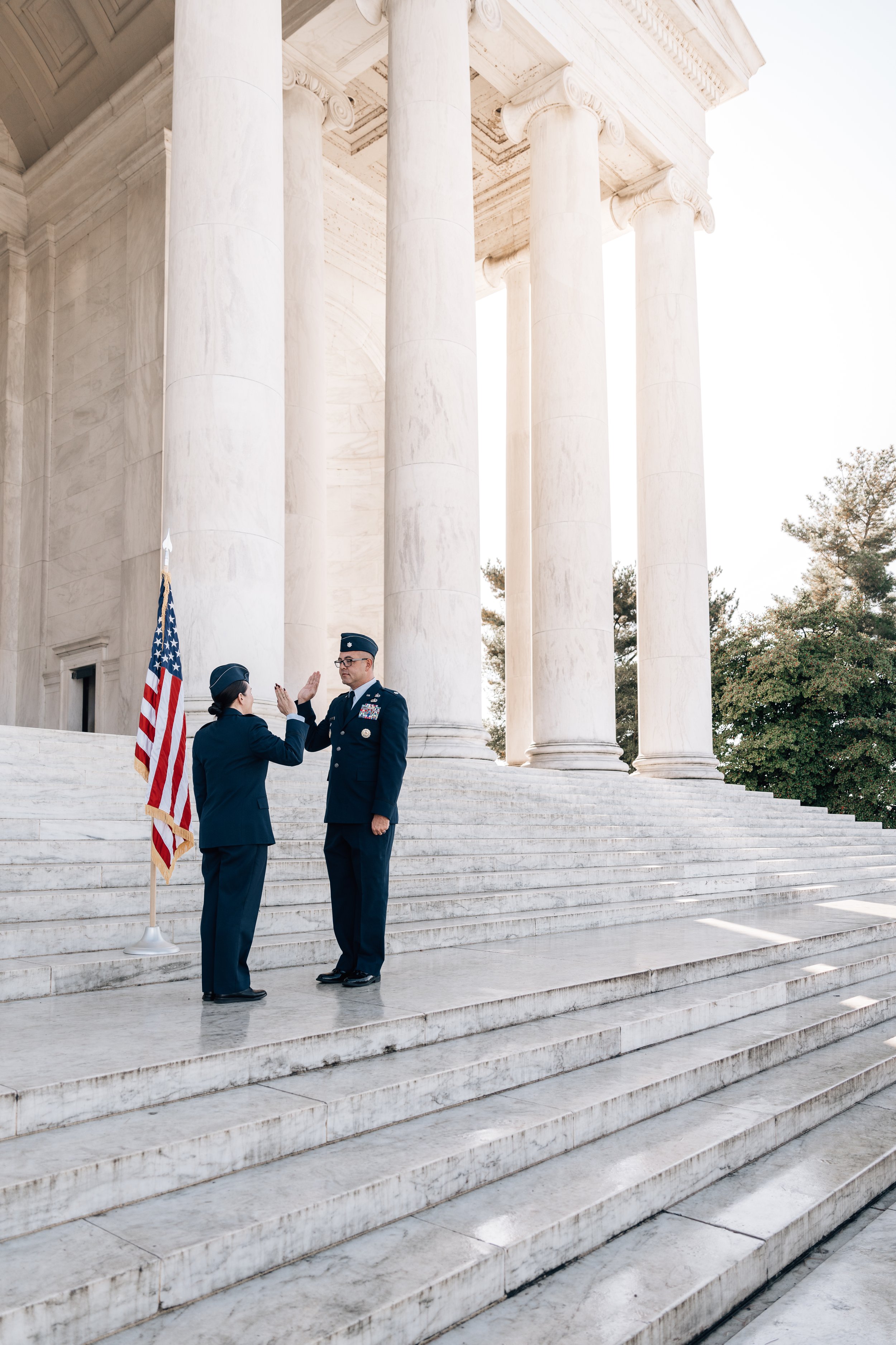 Two uniformed military personnel performing a salute in front of marble columns, American flag beside them, on marble steps.