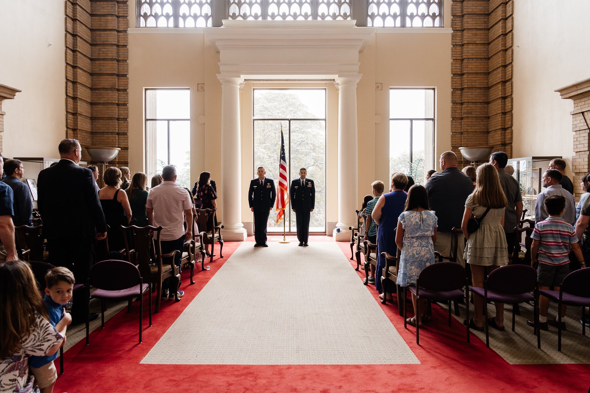 Indoor military ceremony with two officers in uniform standing before an American flag, surrounded by people on a red carpet.