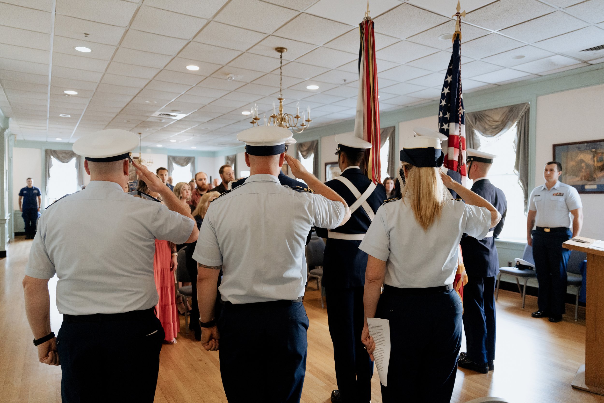Coast Guard personnel saluting during a formal ceremony with a flag display and attendees seated in the background.