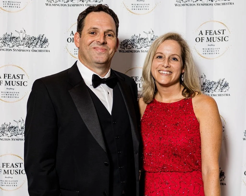 A man in a black tuxedo and a woman in a red dress pose together in front of a Wilmington Symphony Orchestra backdrop.