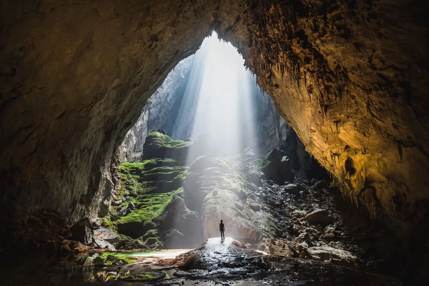A person standing inside a large cave with sunlight streaming down from an opening above, green moss on rocks, and a misty atmosphere.