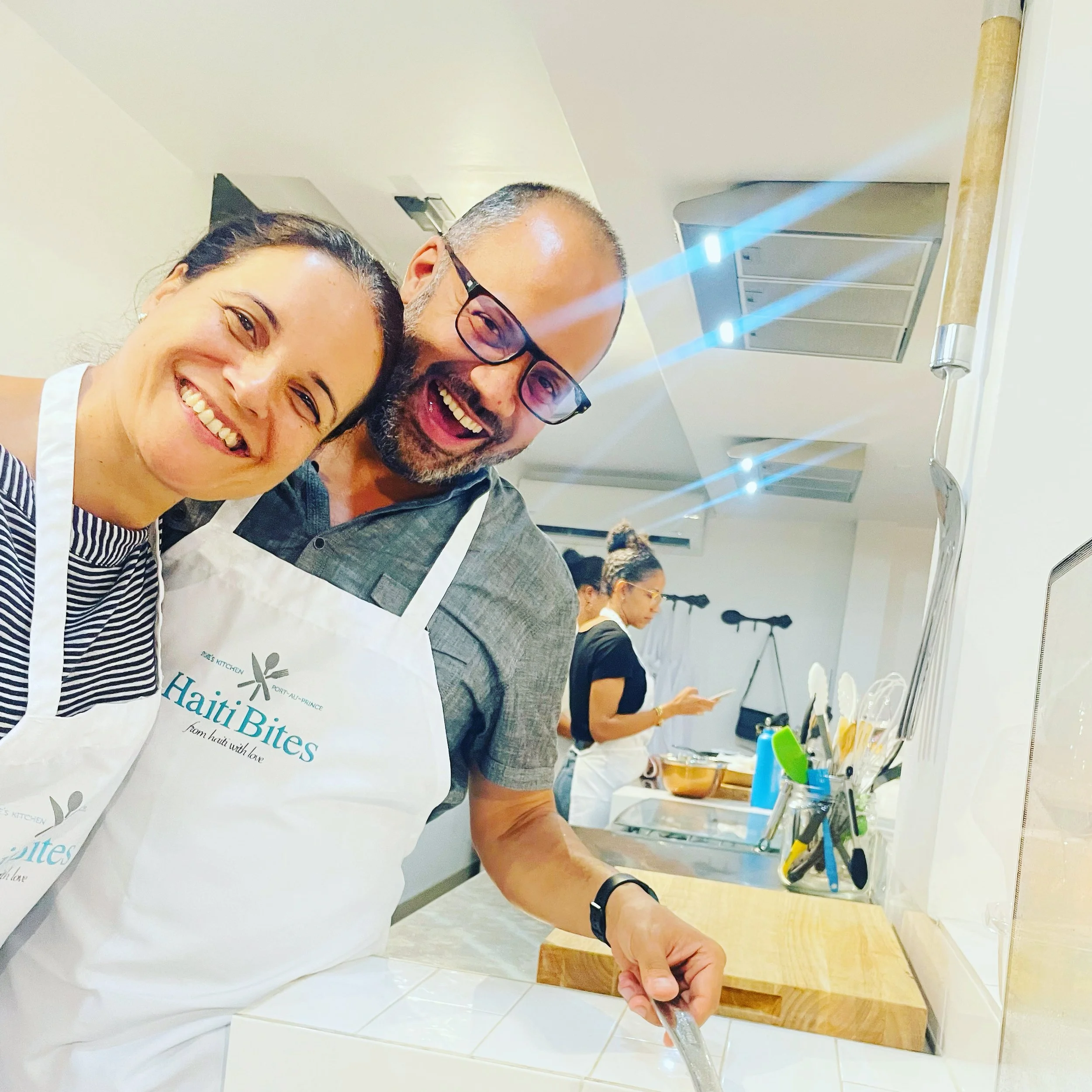 Two people wearing aprons with "Haiti Bites" logo in a kitchen, smiling and cooking. Cooking utensils and another person in the background.