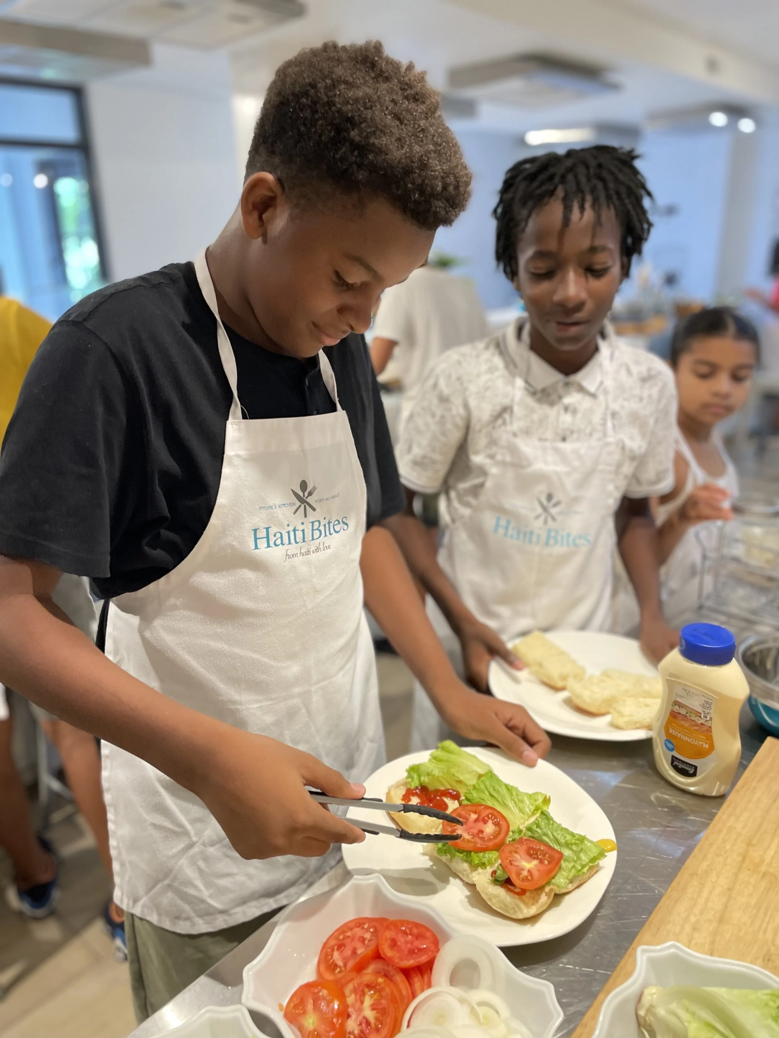 Kids wearing aprons making sandwiches, placing lettuce and tomatoes on bread.