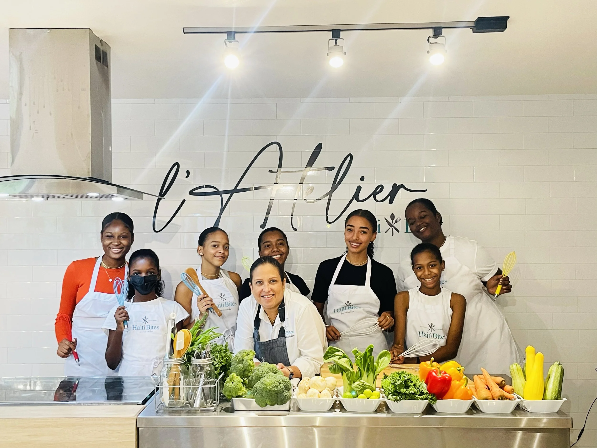 Group of young people in a kitchen setting, wearing aprons and posing with fresh vegetables, under a sign that reads 'l'Atelier'. The kitchen counter displays an assortment of vegetables including broccoli, peppers, and zucchini.