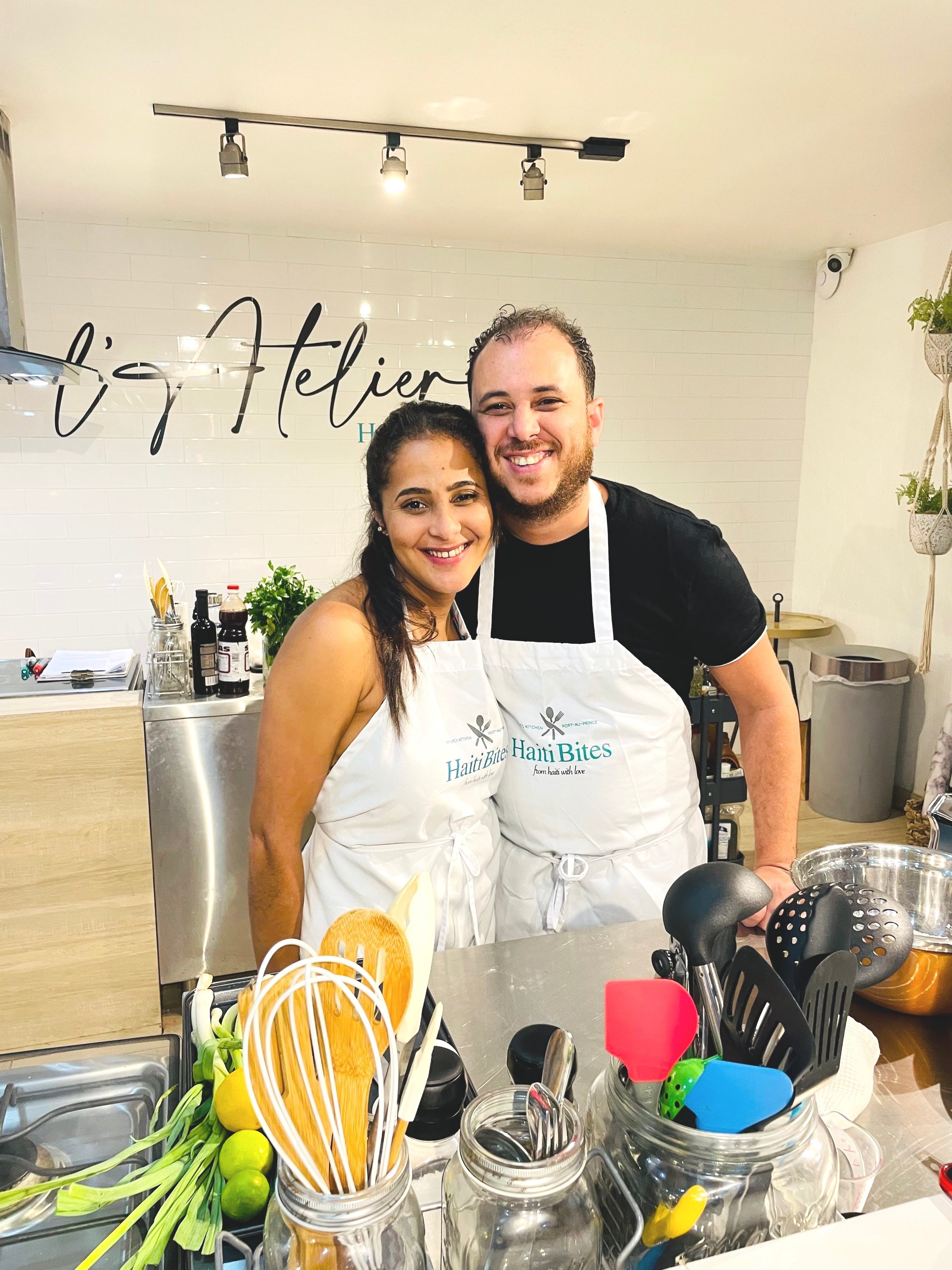 Two people wearing "Haiti Bites" aprons smiling in a kitchen setting with cooking utensils and herbs in the foreground.