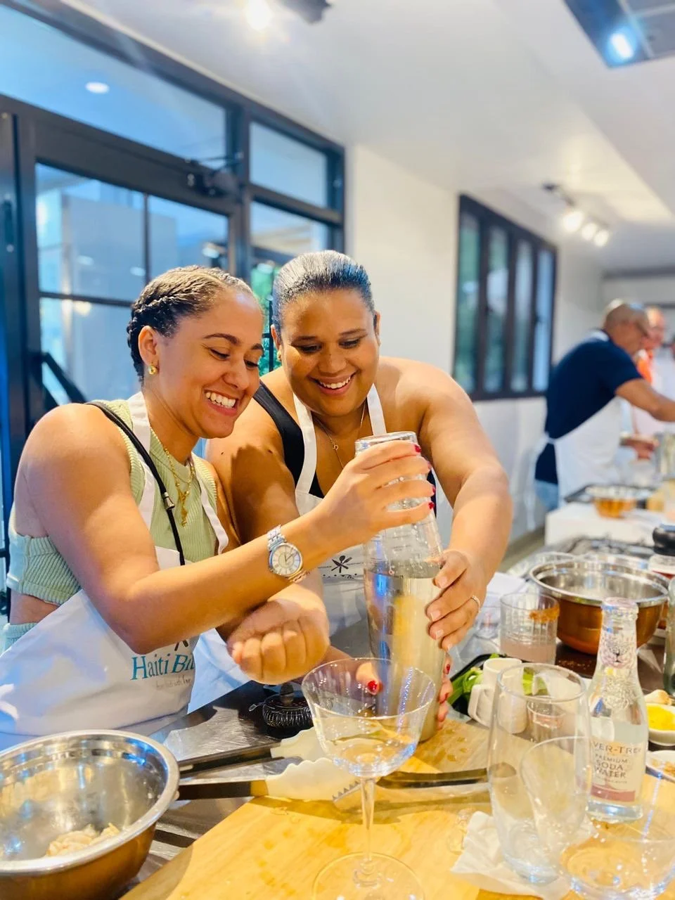 Two women smiling and preparing a drink in a kitchen, wearing aprons, surrounded by kitchen tools, glasses, and ingredients for mixology.