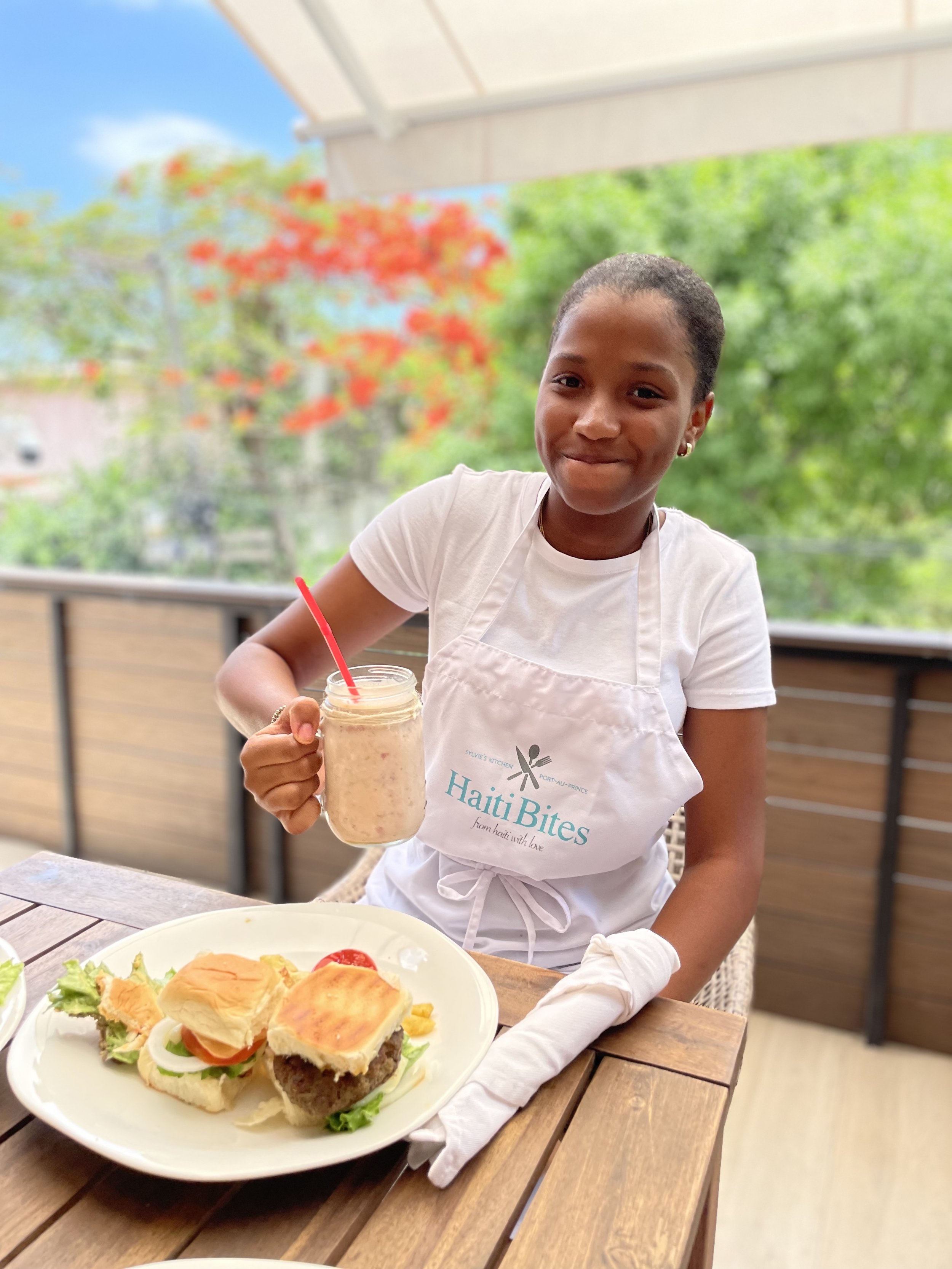 Person wearing "HaitiBites" apron holding a shake and sitting at a table with a plate of burgers, lettuce, tomato, and sauce, with a leafy background.