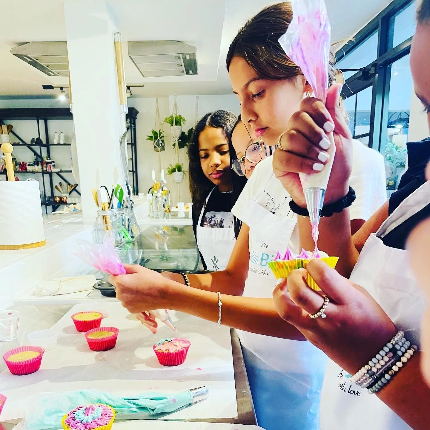 Group of young women decorating cupcakes in a kitchen, using piping bags with colorful frosting, wearing aprons.