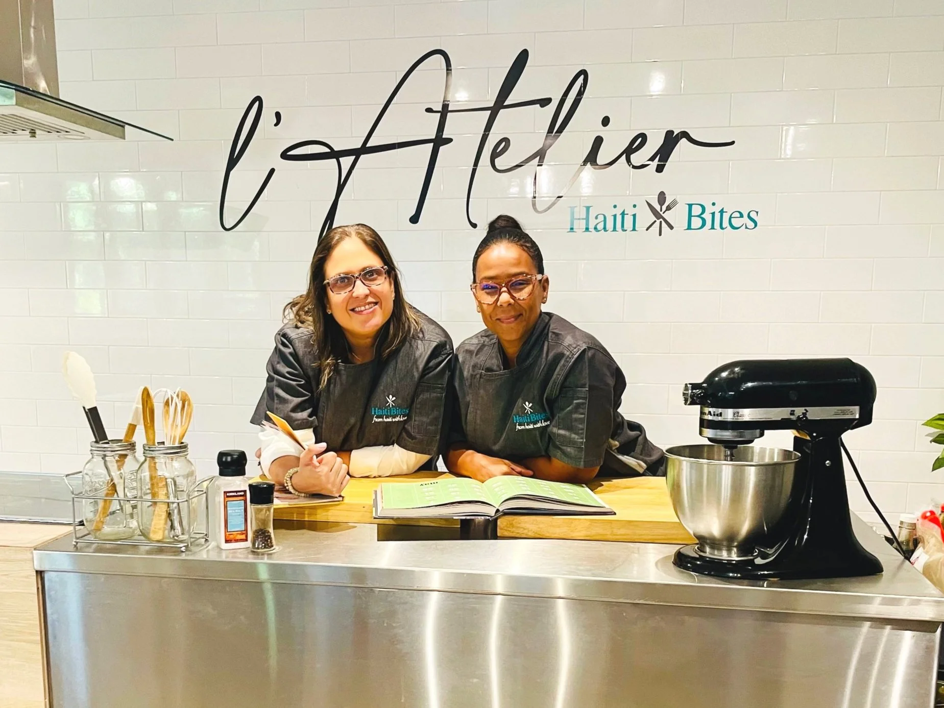Sylvie and Stephanie in chef uniforms at L’Atelier Haiti Bites kitchen preparing food, with kitchen tools and a mixer in the background.