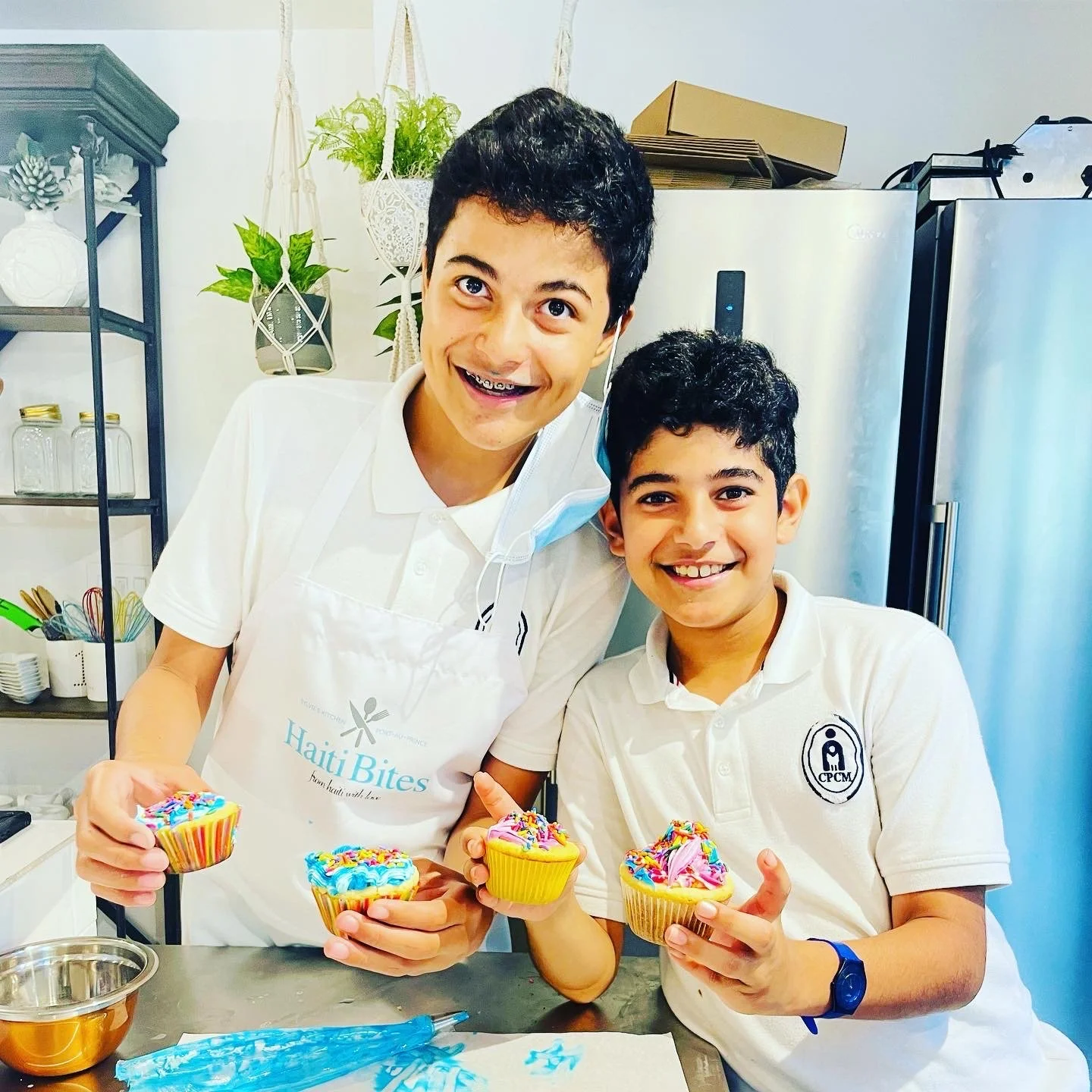 Two children in a kitchen holding cupcakes with colorful sprinkles, wearing white polo shirts, one wearing an apron and a mask under the chin, and smiling.