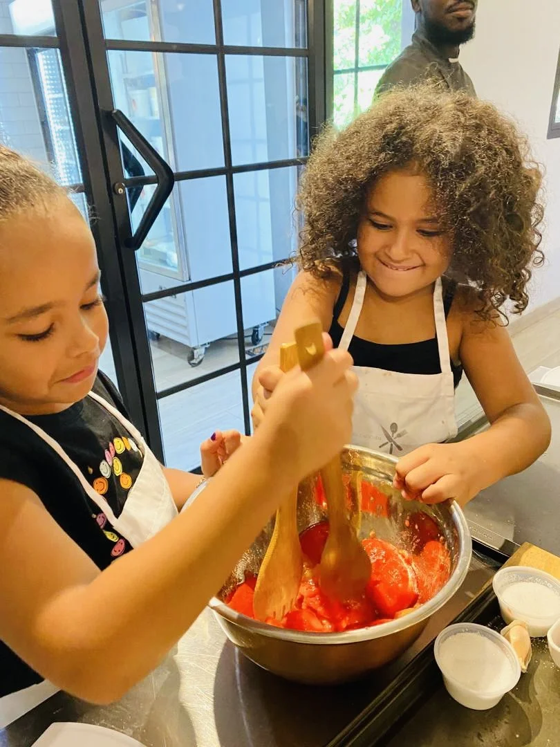 Two children baking, mixing red ingredients in a large bowl using wooden spoons, with a man in the background.