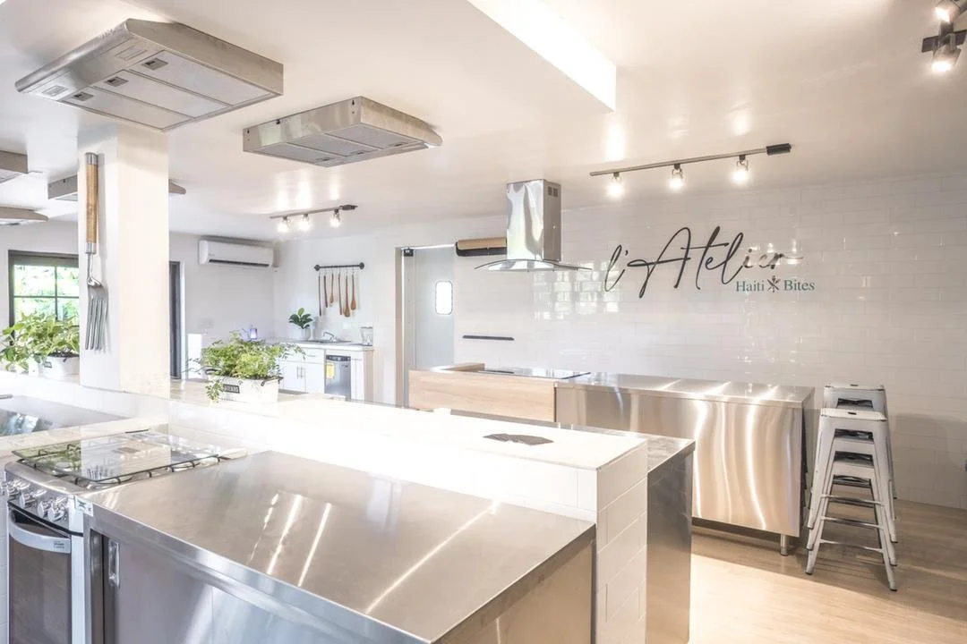 Modern kitchen interior with stainless steel appliances, ceiling vents, plants, utensils, and "l'Atelier Haiti Bites" signage on a tiled wall.