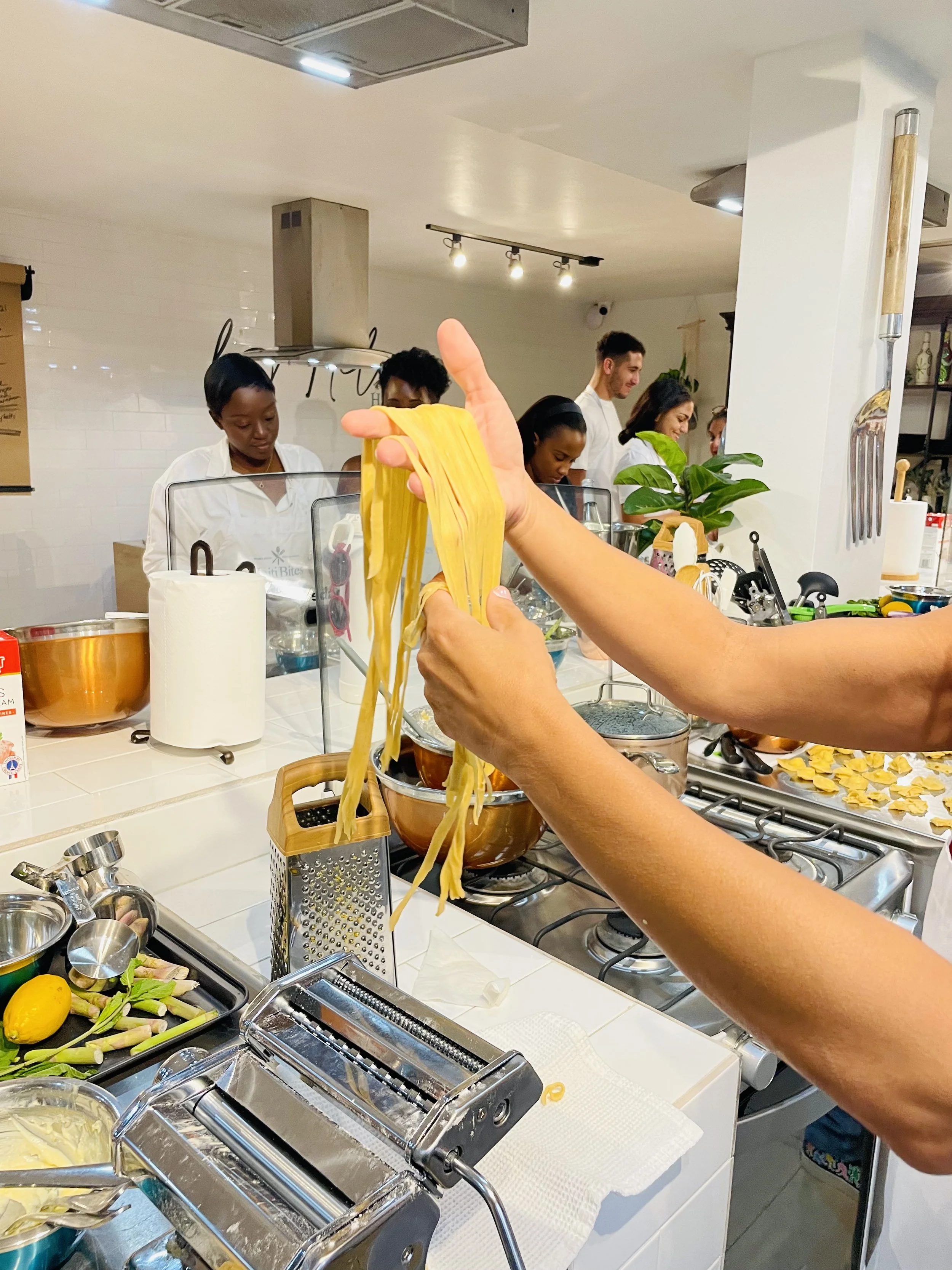 Person handling freshly made pasta ribbons in a kitchen with pasta machine, cooking utensils, and ingredients on the counter.