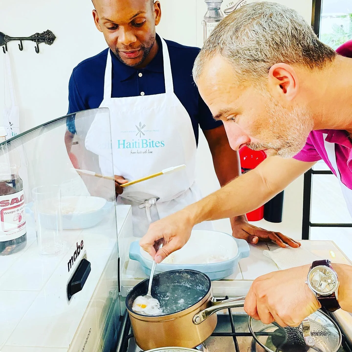 Two men cooking, one stirring a pot on the stove, wearing a white apron and another observing, inside a kitchen setting.